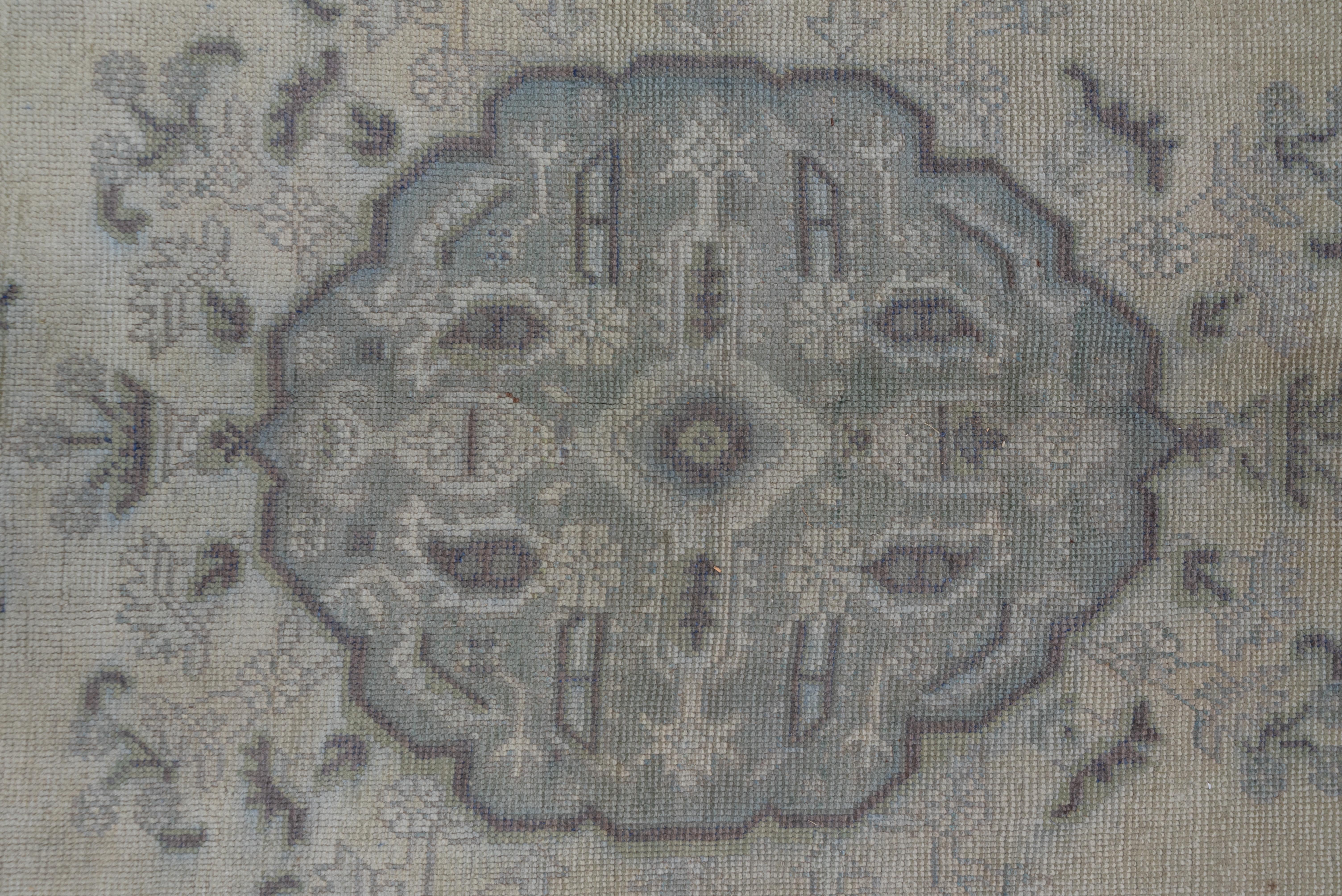 Dieser Werkstattteppich in einem allgemein hellen Farbton aus Sand und hellem Seladon mit Rostakzenten zeigt ein gelapptes Medaillon in einem Blatt- und Blumenkranz sowie große Wappenanhänger. Beide Ecken und die Umrandung weisen Boteh-Blätter auf.