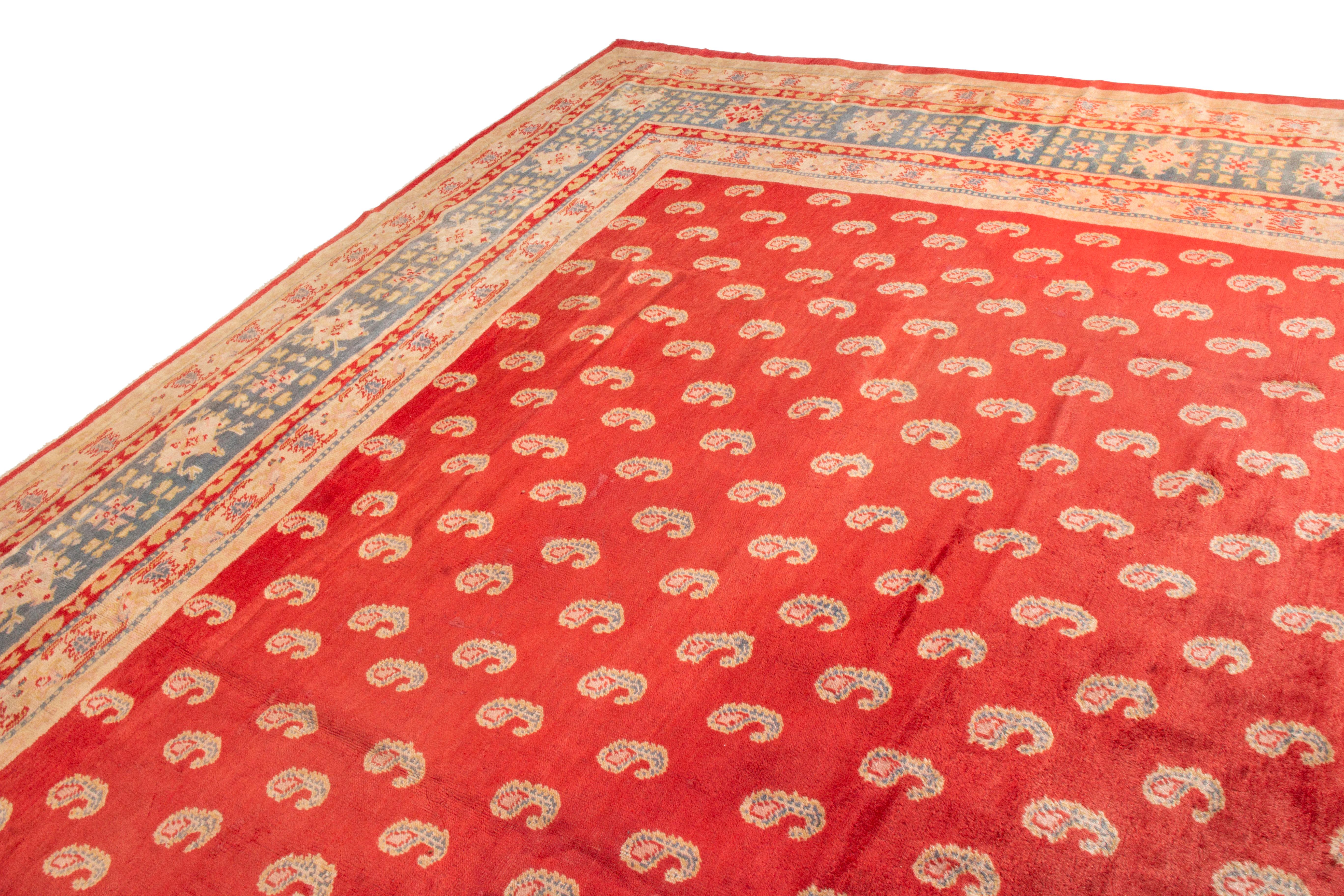 This antique Oushak wool rug has a traditional style with idyllic color ways from this era. Originating from 1910, the combination of bright red, artisan beige, and muted blue is a distinct Oushak trait, though the Paisleys depicted in the field are