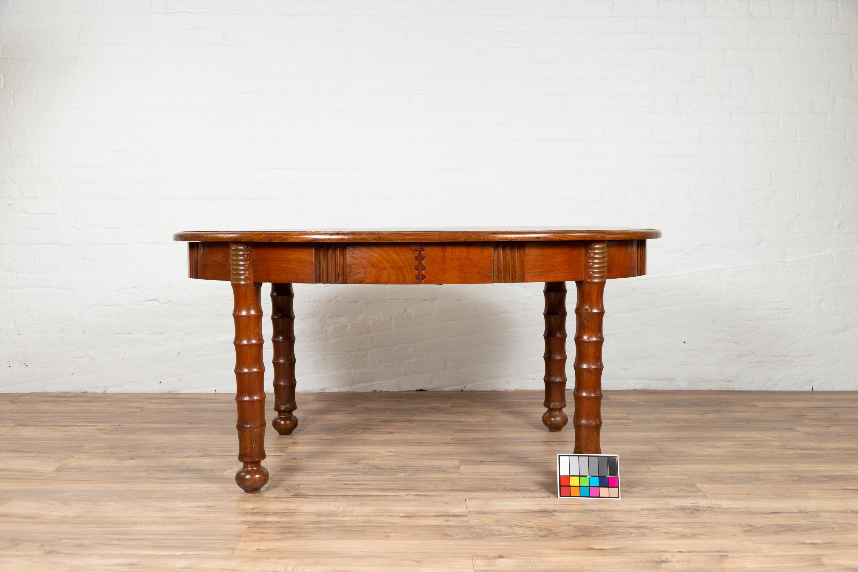 An antique Indonesian wooden oval-shaped dining room table from the early 20th century, with spindle-shaped legs. This elegant Indonesian wooden dining table features an oval top with rounded edges, sitting above an elegant apron accented with