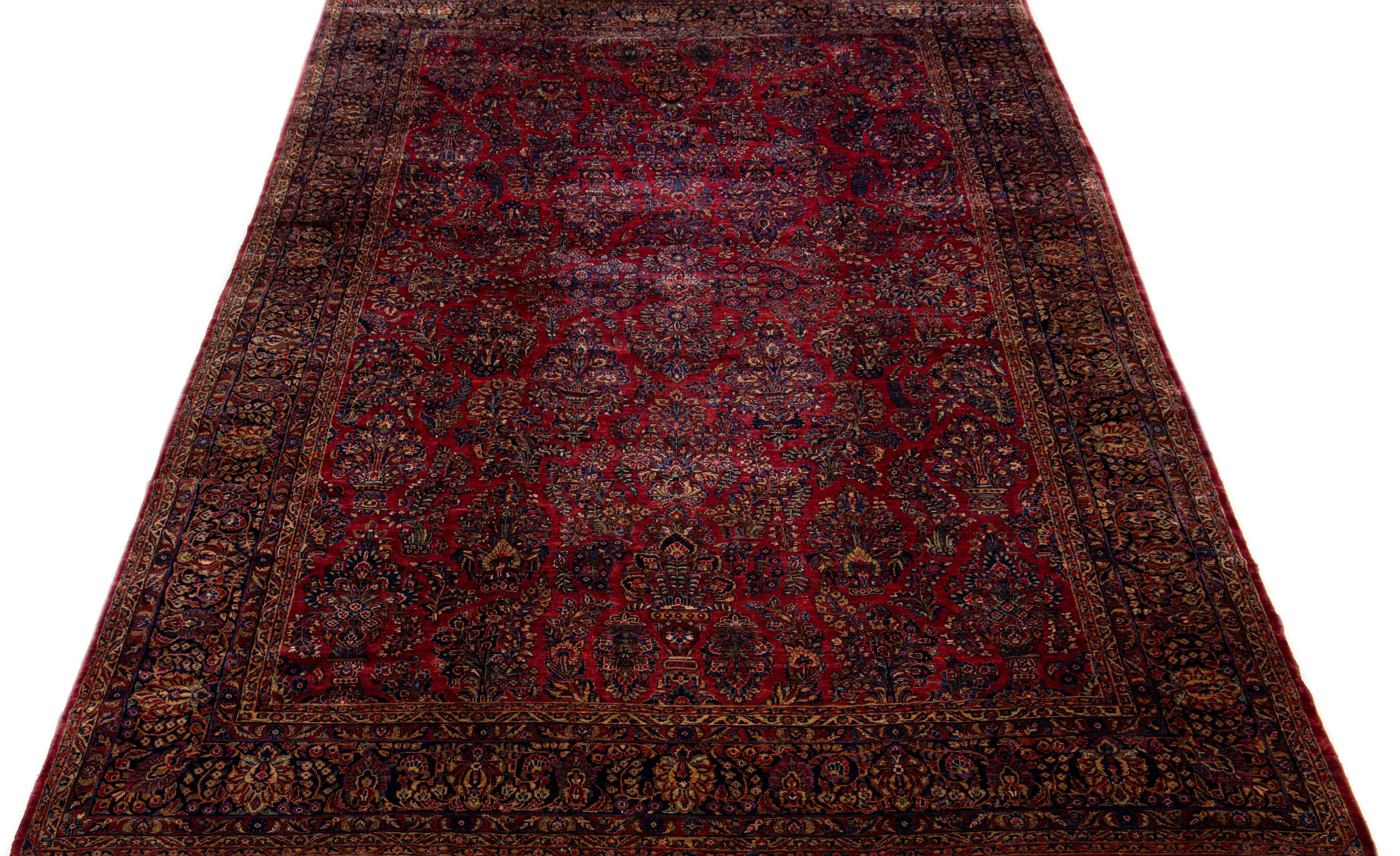 This luxurious antique Persian Sarouk rug is meticulously crafted by hand using fine wool. Its striking red field is elegantly framed by a complex dark blue border adorned with an intricate floral pattern that displays a stunning range of