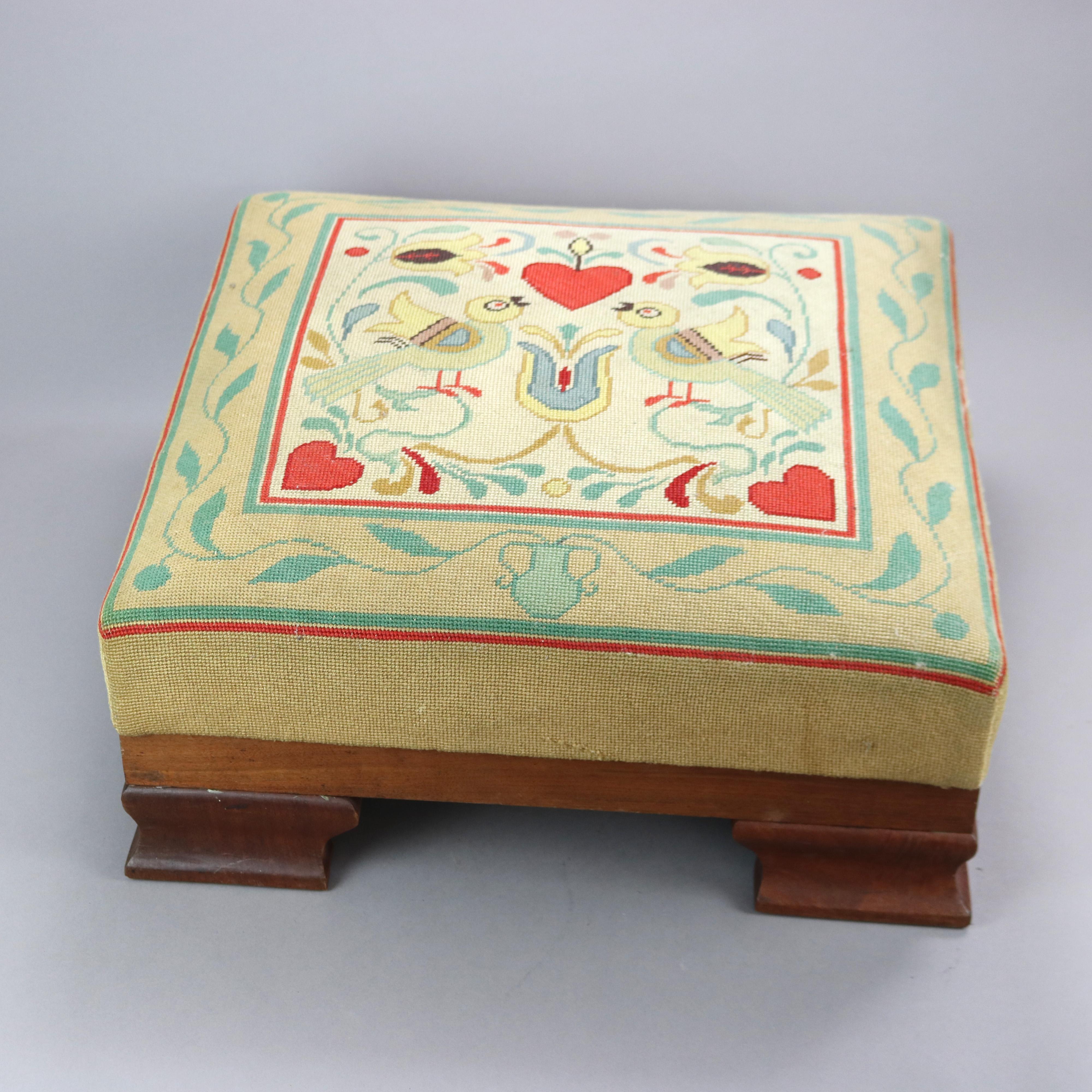 A large antique American Empire footstool offers needlepoint upholstered seat with German Fraktur style design having lovebirds, hearts and flowers over walnut frame raised on ogee bracket feet, c 1890 

Measures - 12''H x 24.5''W x 24.5''D.