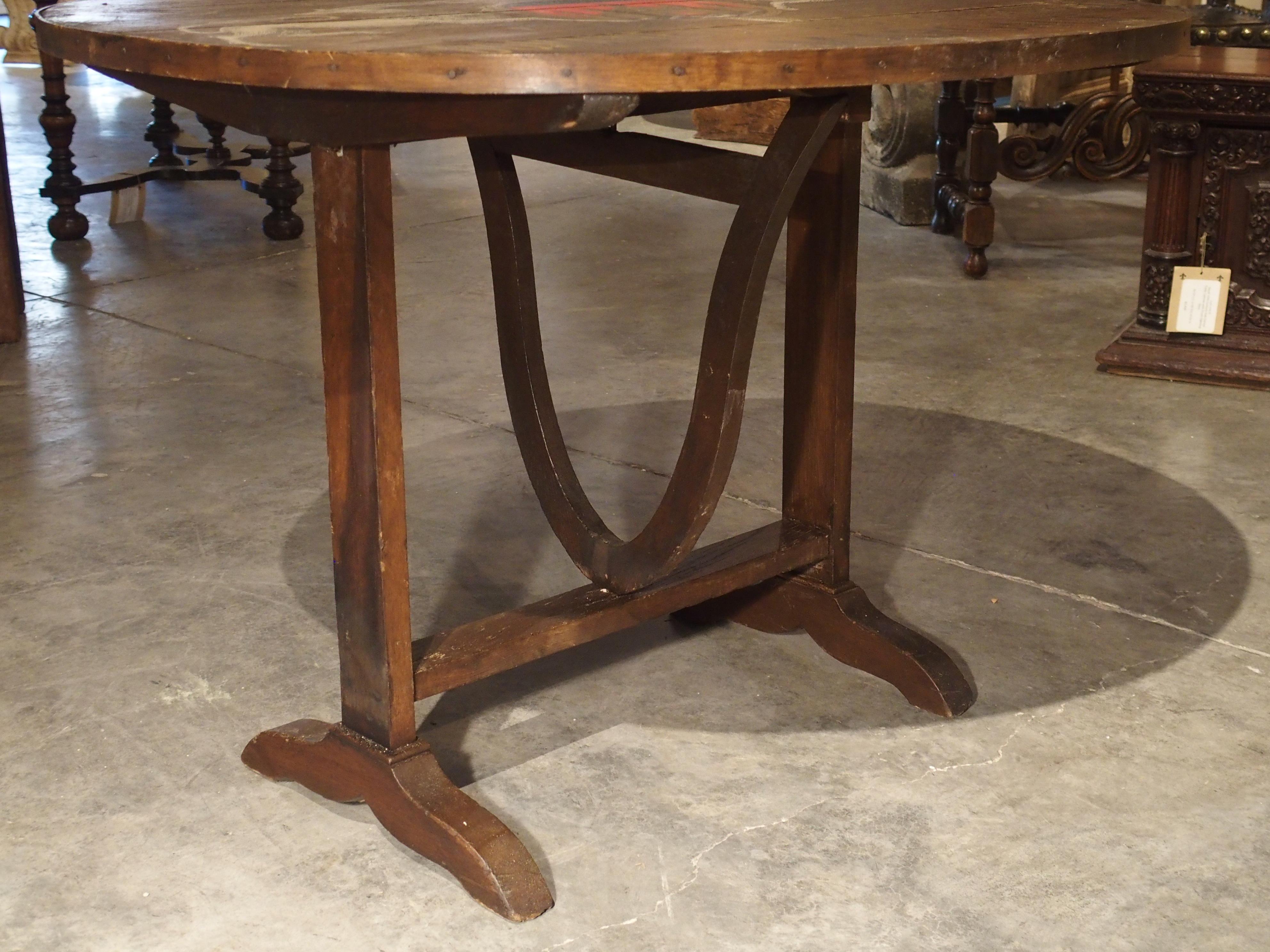 From circa 1900, this wine tasting table is of typical size and has the circular shape of other French tasting tables. However, later in the 20th century, its tilt top was wonderfully painted to depict the colorful crest of the French vineyard, Haut