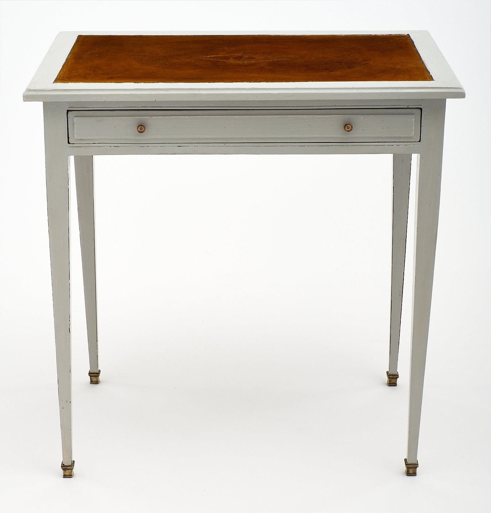 Painted French antique writing desk topped with tan gilt embossed leather in the Louis XVI style. This piece is made of cheerywood and painted Trianon dove-gray. It is a practical piece featuring a large dovetailed drawer and two pull-out leaves