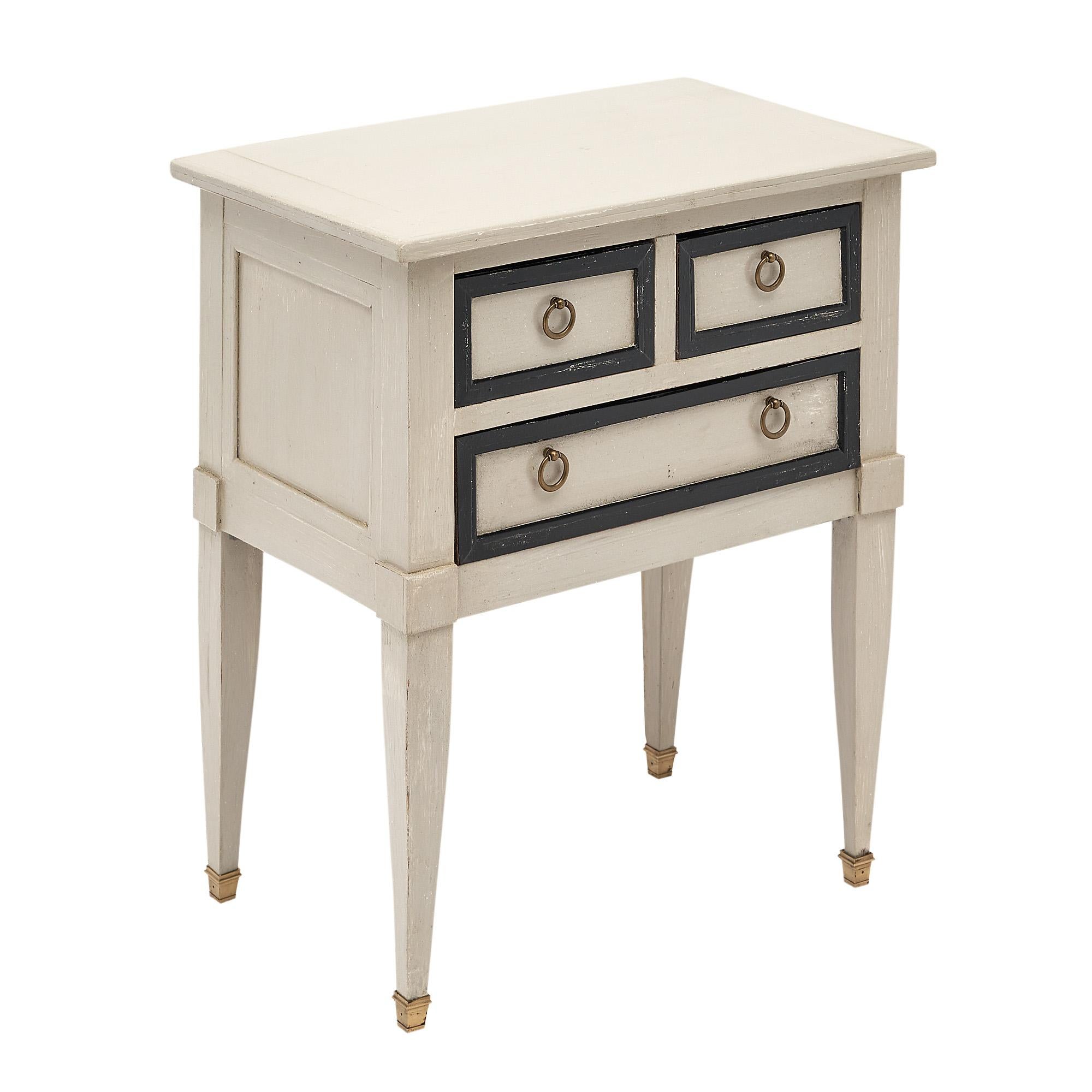 Side table, French, made of cherry wood with a hand painted “Trianon” gray finish and black hand painted trim. Three dovetailed drawers have brass hardware rings. The tapered legs are capped with brass feet.