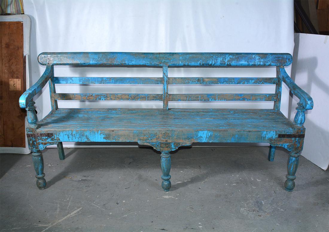 The wonderfully blue-painted Dutch Colonial style solid teak wood bench has slatted back and slightly concave seat for comfort. Metal straps secure all four corners. The front legs and arm supports are turned. Benches like these were used in the