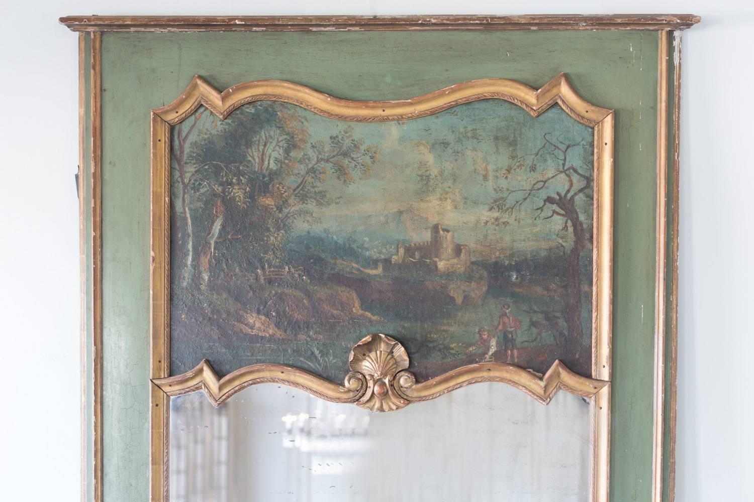 Stunning trumeau mirror in dark forest green with a painting depicting a castle, river, and two boys in the foreground. The beautiful image is framed by a contrasting gilt leading to a central medallion. This stately and alluring piece does show a