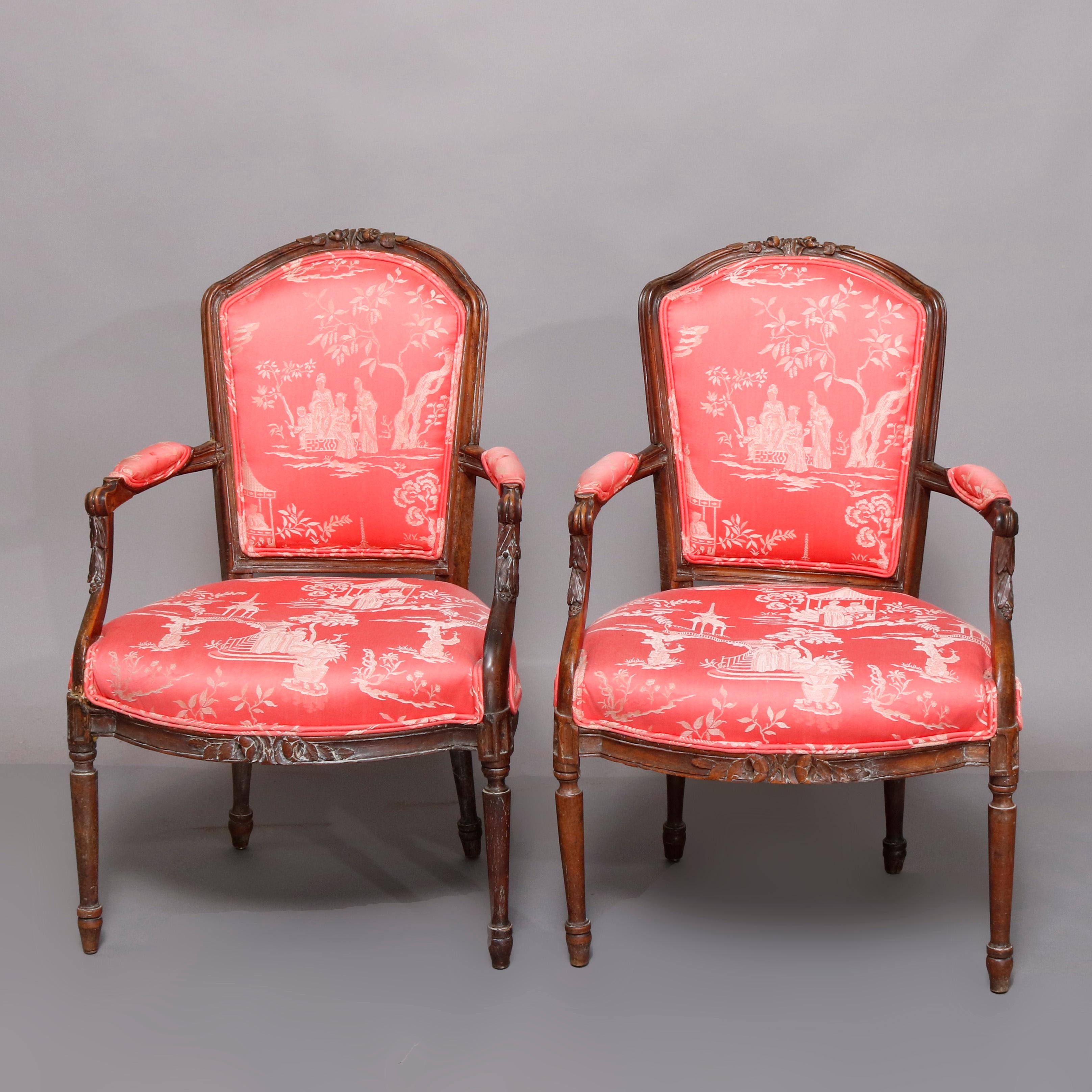 An antique pair of French Louis XVI armchairs offer carved fruitwood frames having foliate and floral crests surmounting shield form backs and with covered scroll form arms terminating in carved foliate supports and raised on turned and tapered