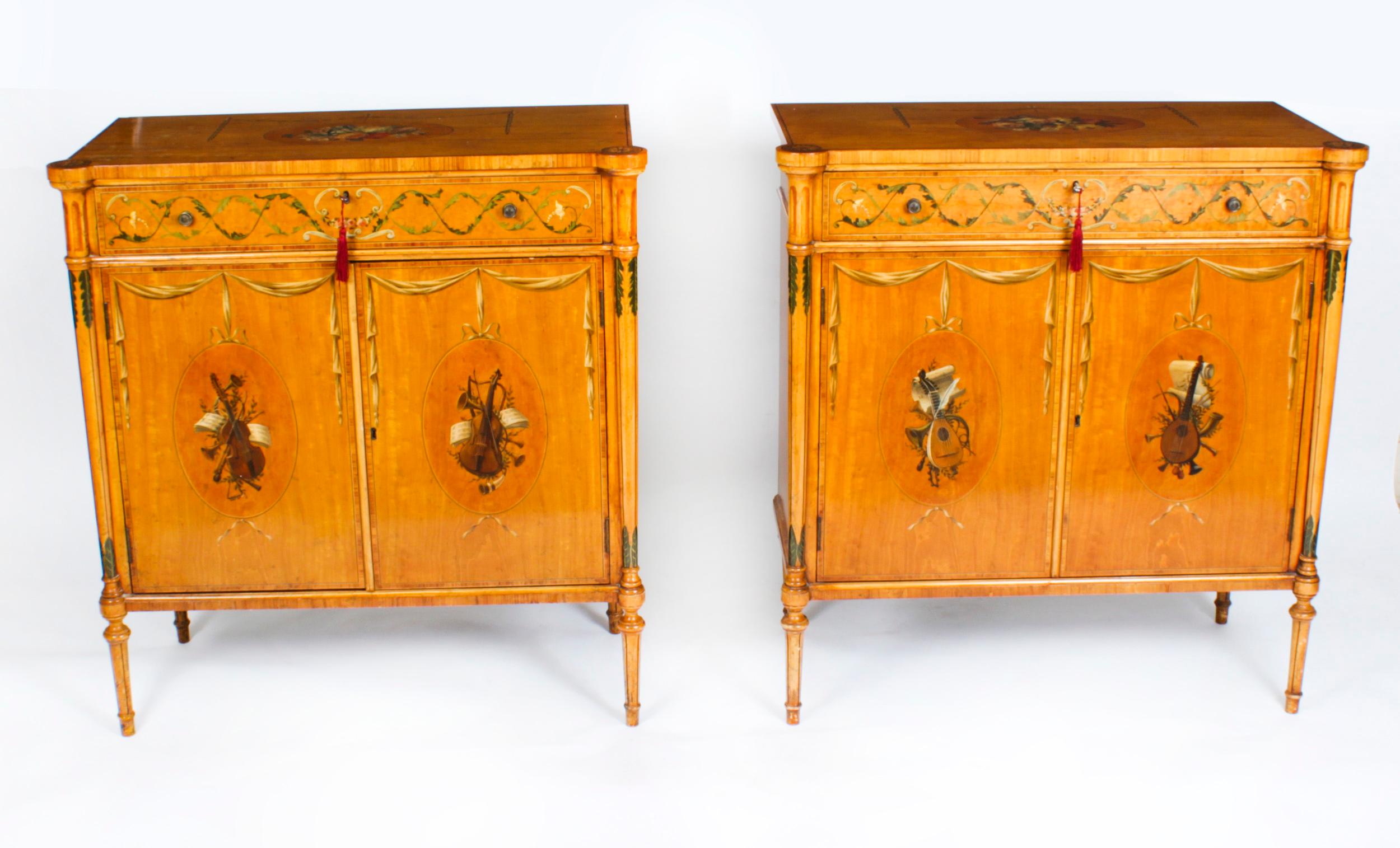 This is a gorgeous antique pair of Adam Revival two door satinwood side cabinets with beautiful hand painted floral decoration in the manner of Angelica Kauffman, dating from the late 19th century.
 
The tops beautifully decorated with a bouquet