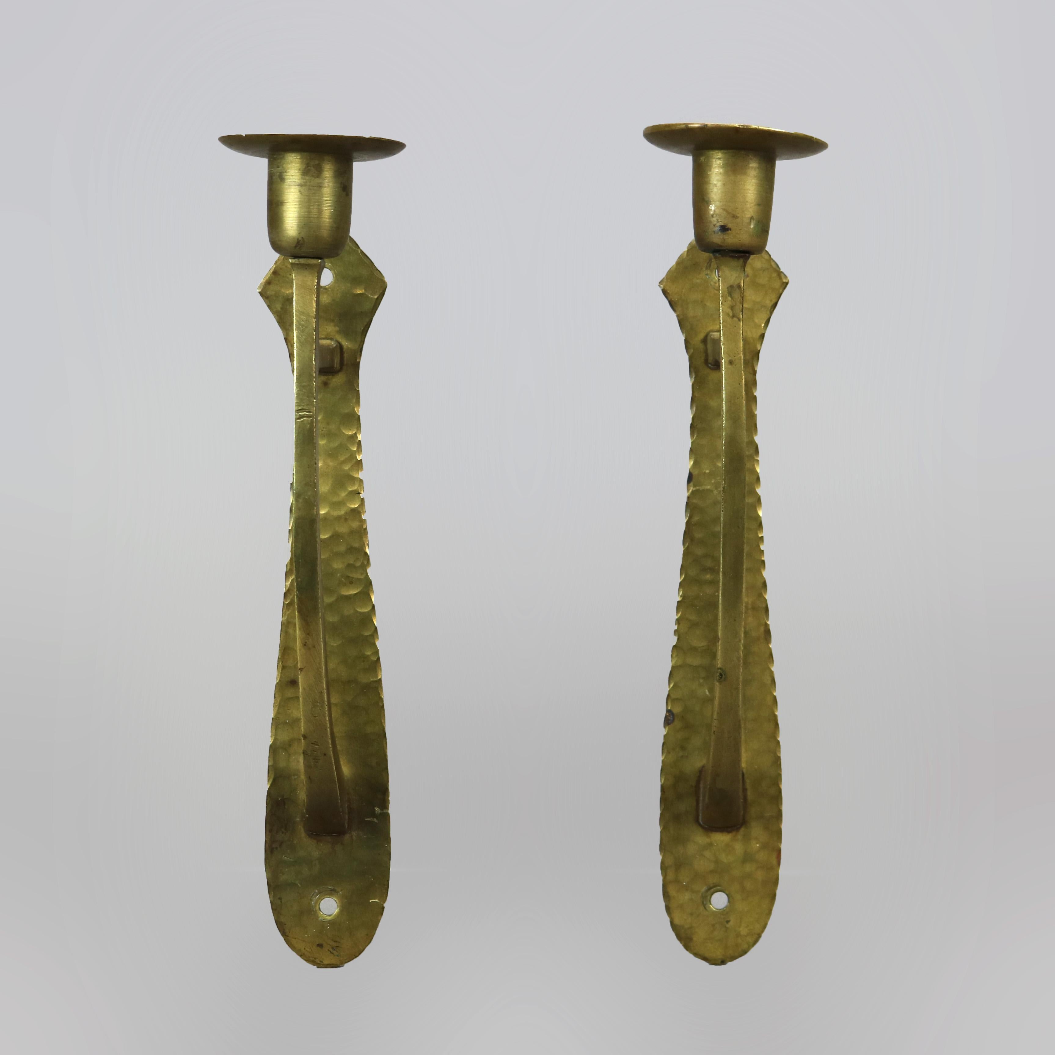 A pair of antique Arts & Crafts wall sconces by Gustav Stickley offers hammered brass construction with stylized shield form plate having scroll arm terminating in candle socket, guaranteed by Gustav Stickley and referenced in the catalog,