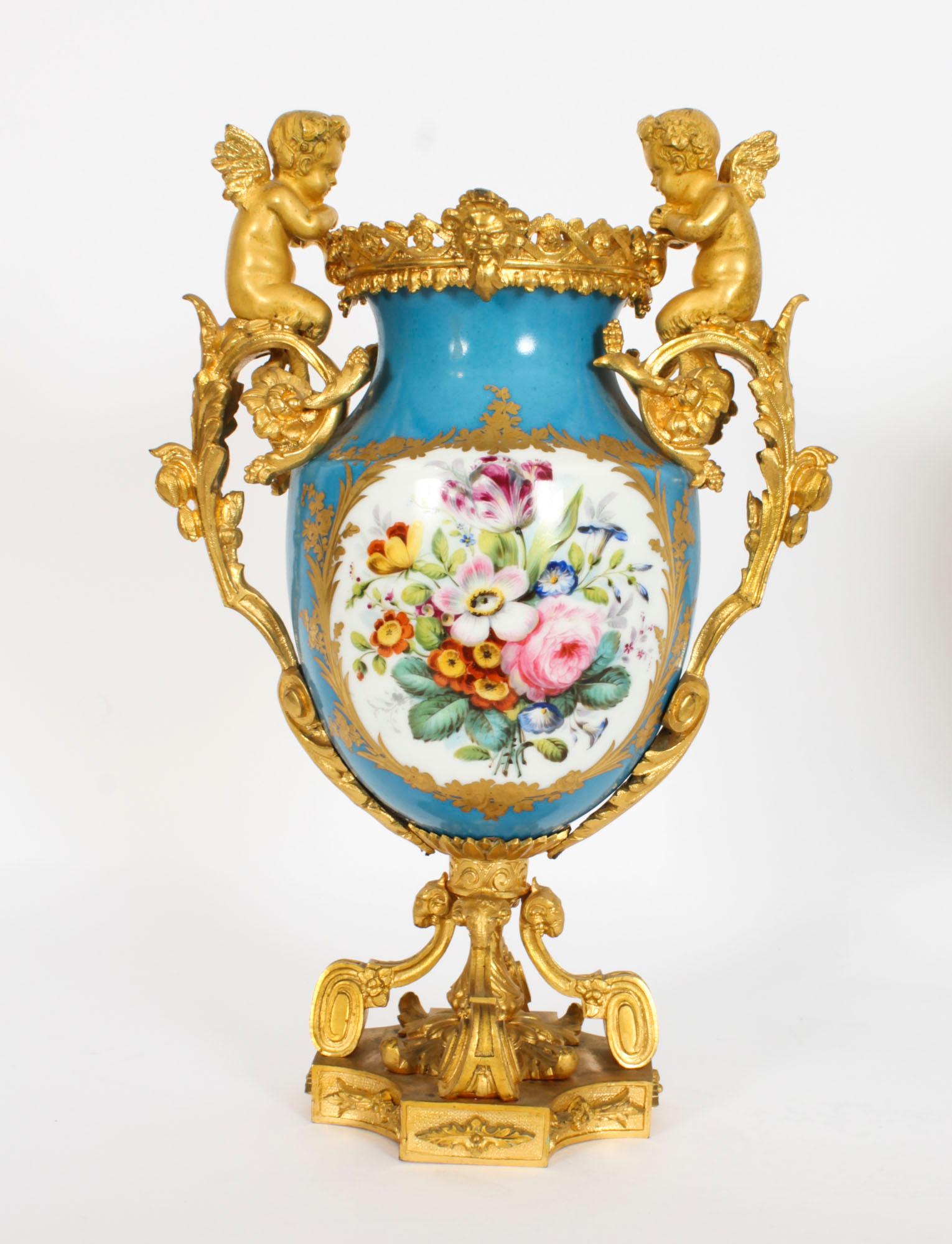 This is a stunning pair of French twin handle ormolu garniture urns with Sevres hand painted porcelain, circa 1870 in date.

The urns are decorated in the Sevres manner with panels of cherubs and bouquets of flowers reserved on a blue ground, the