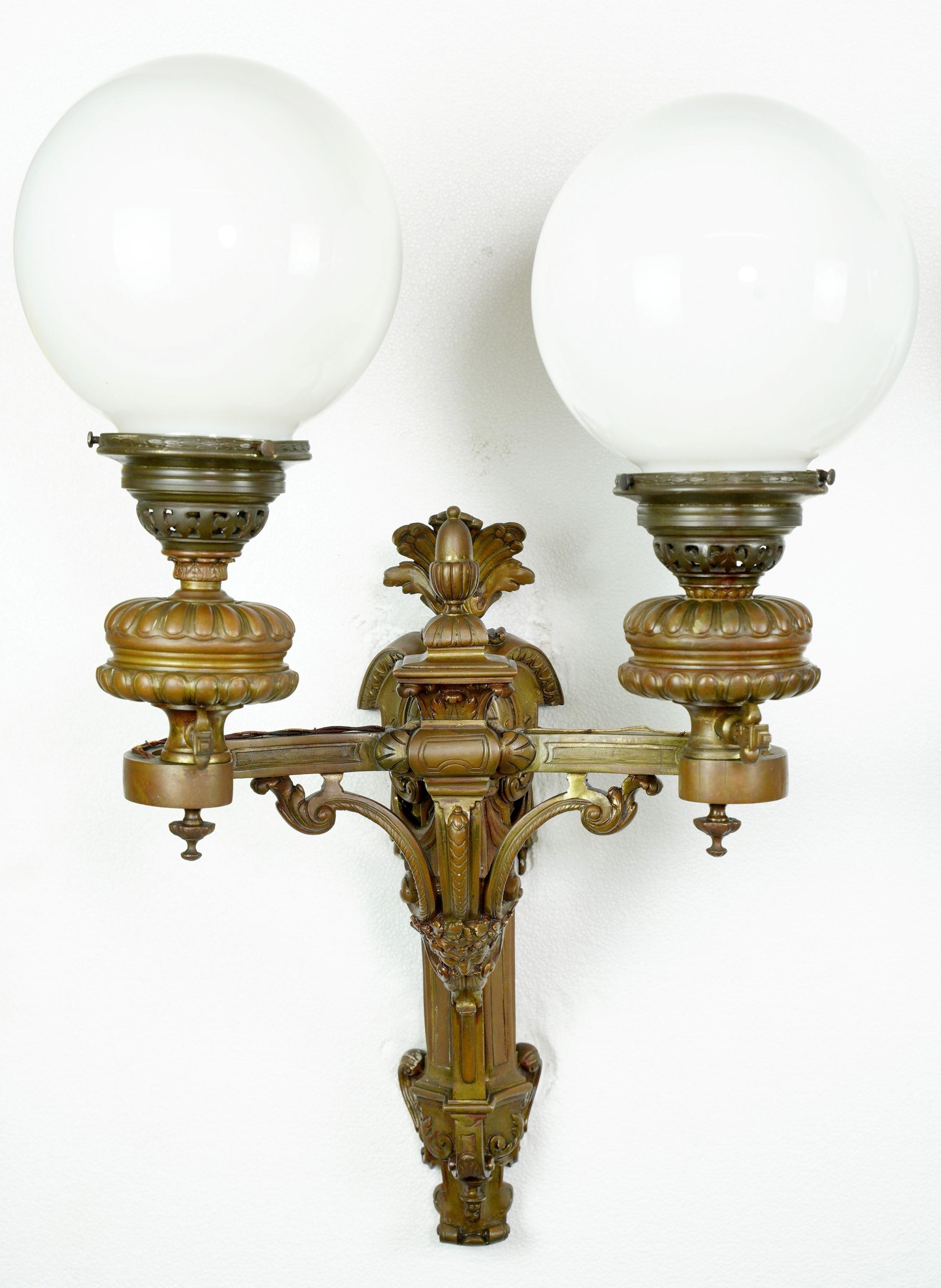 Early 20th Century pair of bronze antique two arm sconces with milk glass spherical globe shades and featuring standard sockets. Cleaned and restored. One of the two sconces has a slightly different lower base. Please see images. Priced as pair.