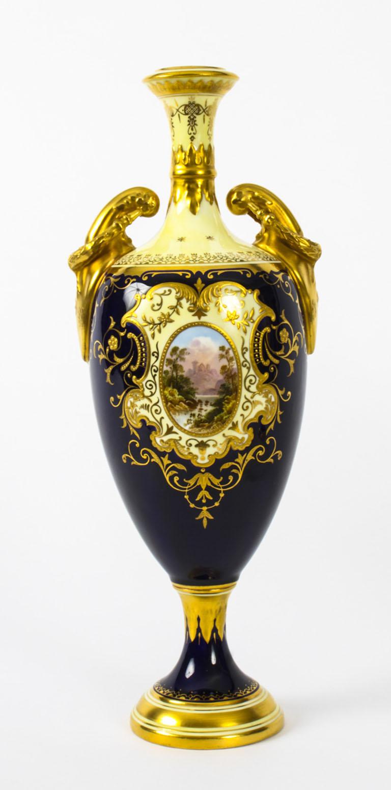 A superb pair of Coalport pedestal ovoid vases, late 19th century in date.

The painted oval panels feature handpainted landscapes set within shaped cartouches on a cobalt blue ground with lemon borders and gilt flying scroll handles. Both are