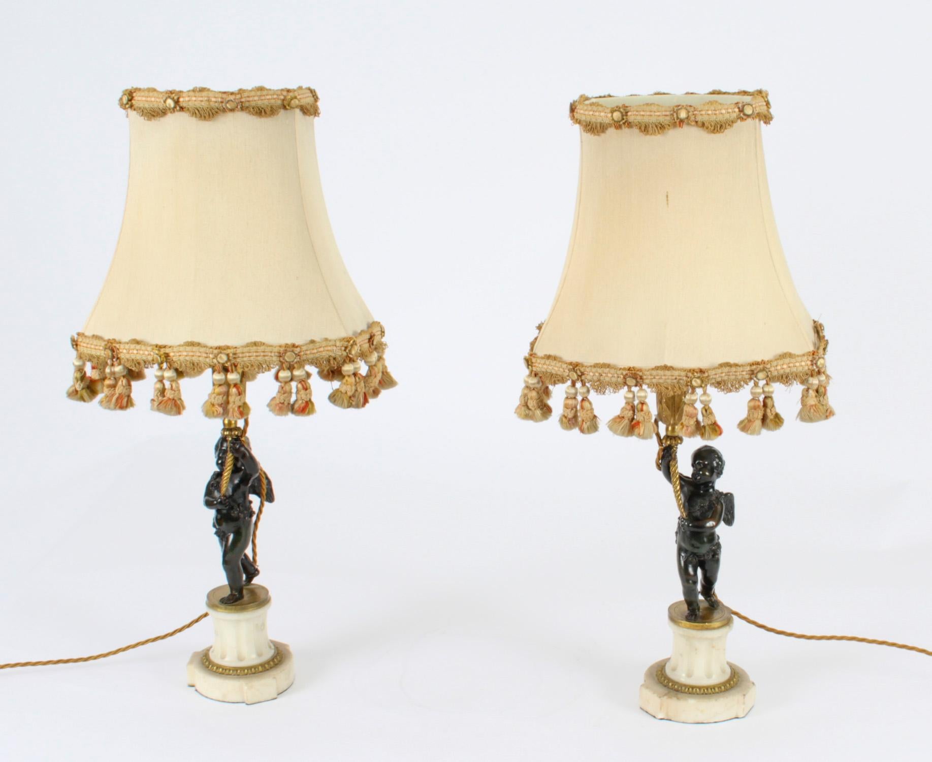 A magnificent antique pair of French Louis Revival patinated and parcel gilt bronze and marble mounted figural table lamps with shades, late 19th century in date.

Each modelled with a cherub raising a foliate cast light fitment, and raised on