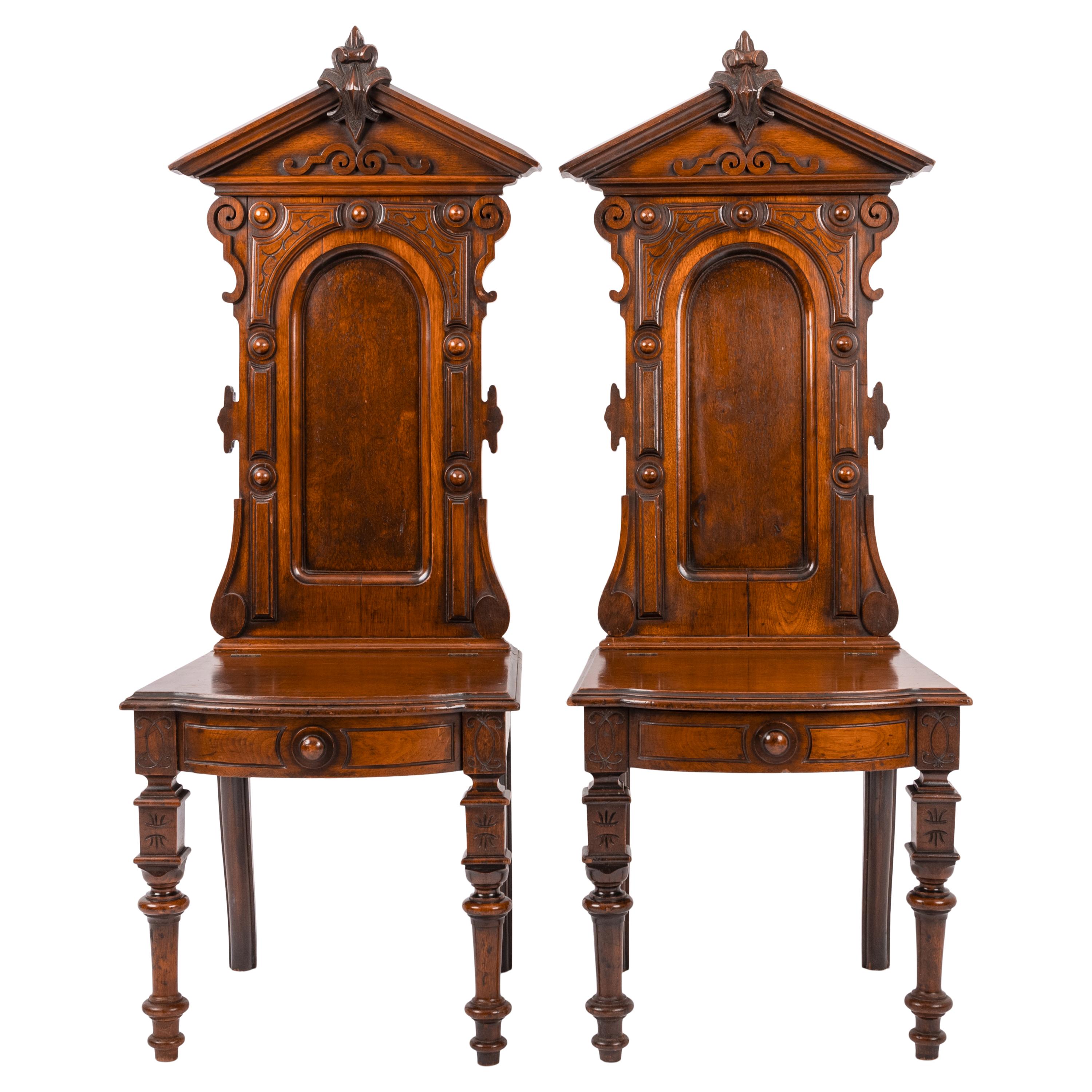 A good pair of antique American Renaissance revival carved walnut hall chairs, circa 1870.
The chair with high backs and having a neo-classical architectural pediment with a carved fleur-de-lis crest, to the center is a burl walnut arched panel,