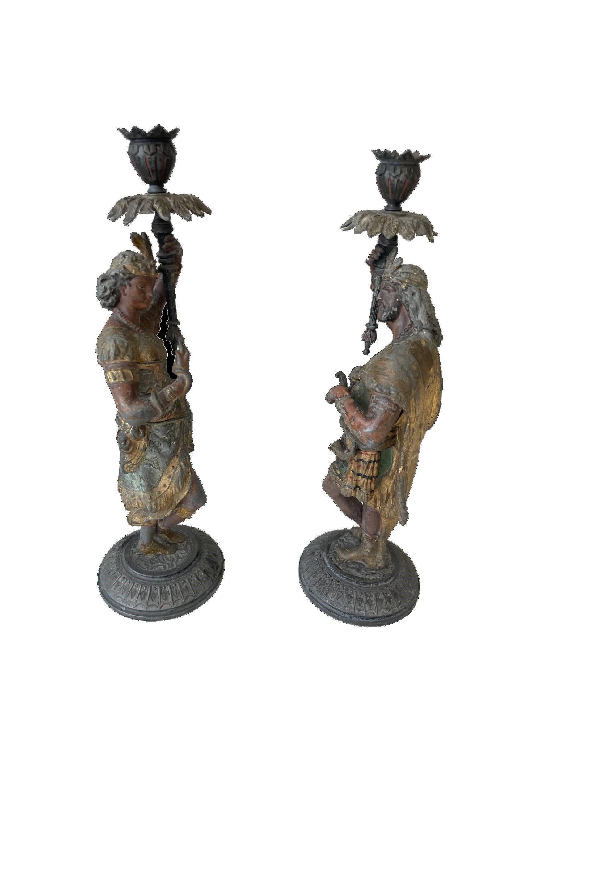 A strikingly impressive pair of figural candle holders, cast in the form of finely modelled Regal Indian male & female figures. Each standing atop circular bases and holding aloft floral shaped vessels serving as candle nozzles. Decorated overall