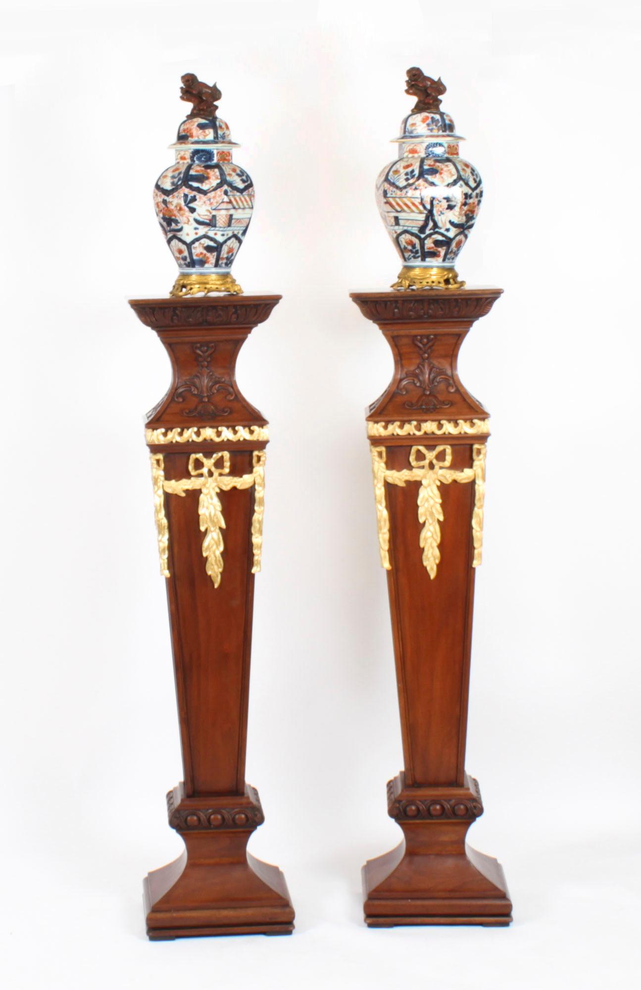 This is a gorgeous pair of Antique English Sheraton Revival design mahogany and gilt wood pedestals, C1920 in date. 
 
The square tops above anthemion carved concave friezes with floral gilt swags below, the tapering panelled columns standing on