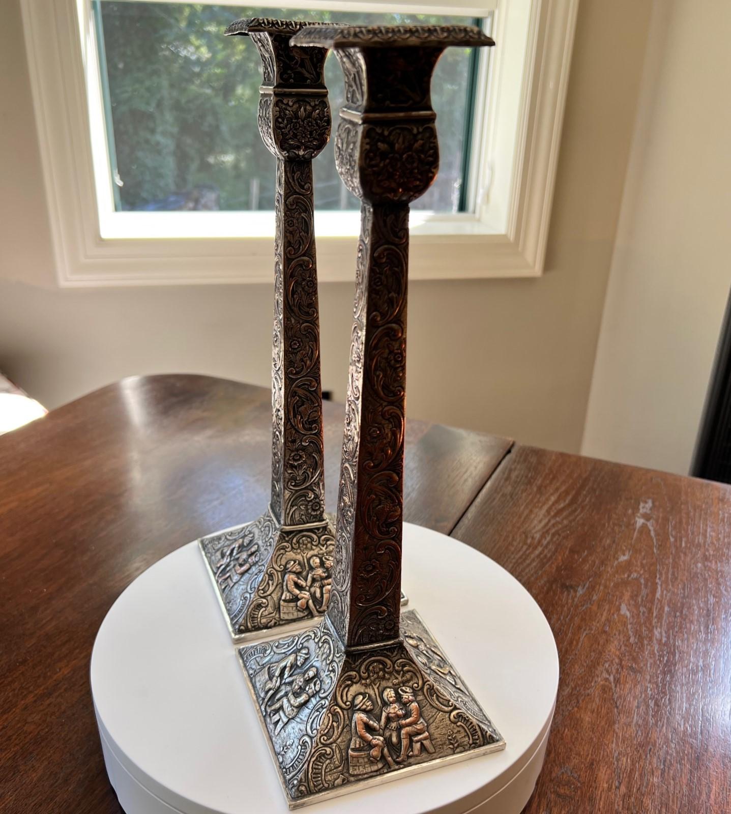 E.G. Webster and Sons silver plate repousse candlesticks, each with squared bobeche decorated with birds, over the floral standard, rising on a square base with Dutch tavern scenes, marked underfoot.

Dimensions:
13.5