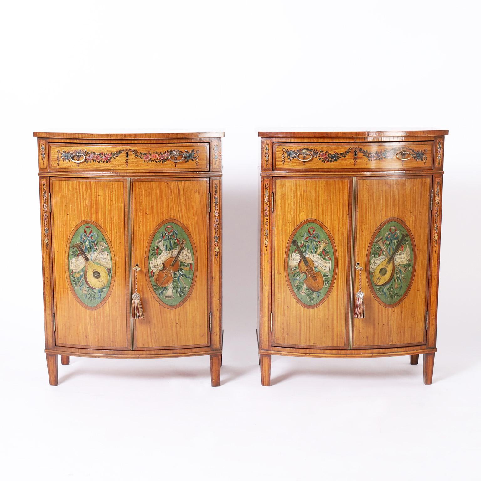 Standout pair of antique English bow front cabinets crafted in satinwood and mahogany with one drawer and two doors paint decorated with musical motifs such as horns, violins and lutes in classic Adam style.