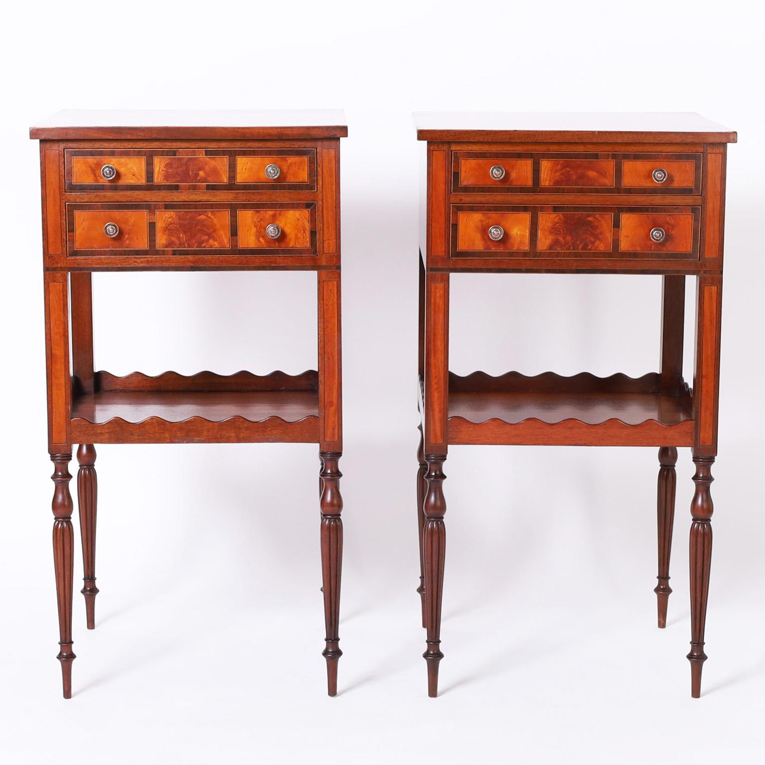 Handsome pair of antique Sheraton style English stands crafted in mahogany with a crossbanded top on a case with two drawers featuring crossbanded and burled panels and brass hardware. The lower tier has a scalloped gallery and sits on elegant