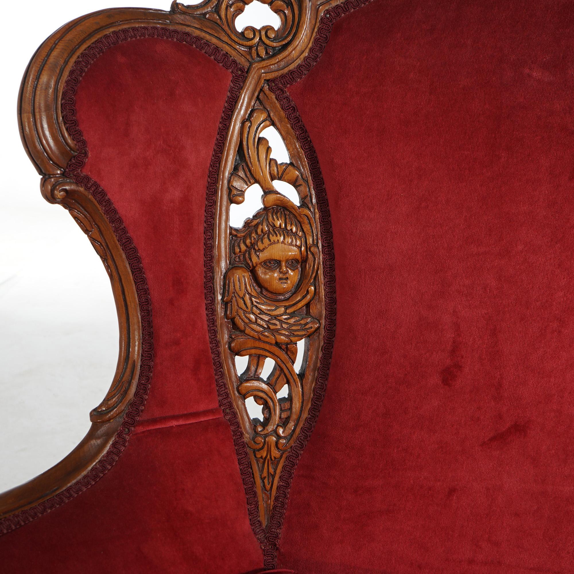 An antique pair of French figural fireside wingback chairs offers walnut construction with crest having carved birds over upholstered backs and seats with carved cherub and foliate elements, raised on cabriole legs terminating in scroll form feet,