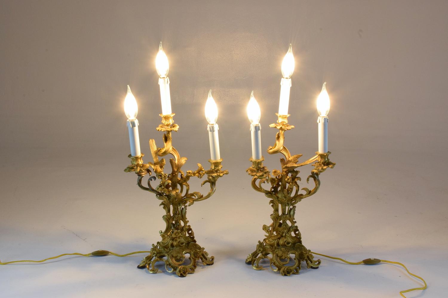 A pair of french antique mid-19th century Louis VXI Rocaille style electrified three-arm candelabra lamps entirely mounted in ormolu bronze doré. The ormolu is meticulously crafted with a beautifully gilt foliage of s scrolls, leafs, birds and