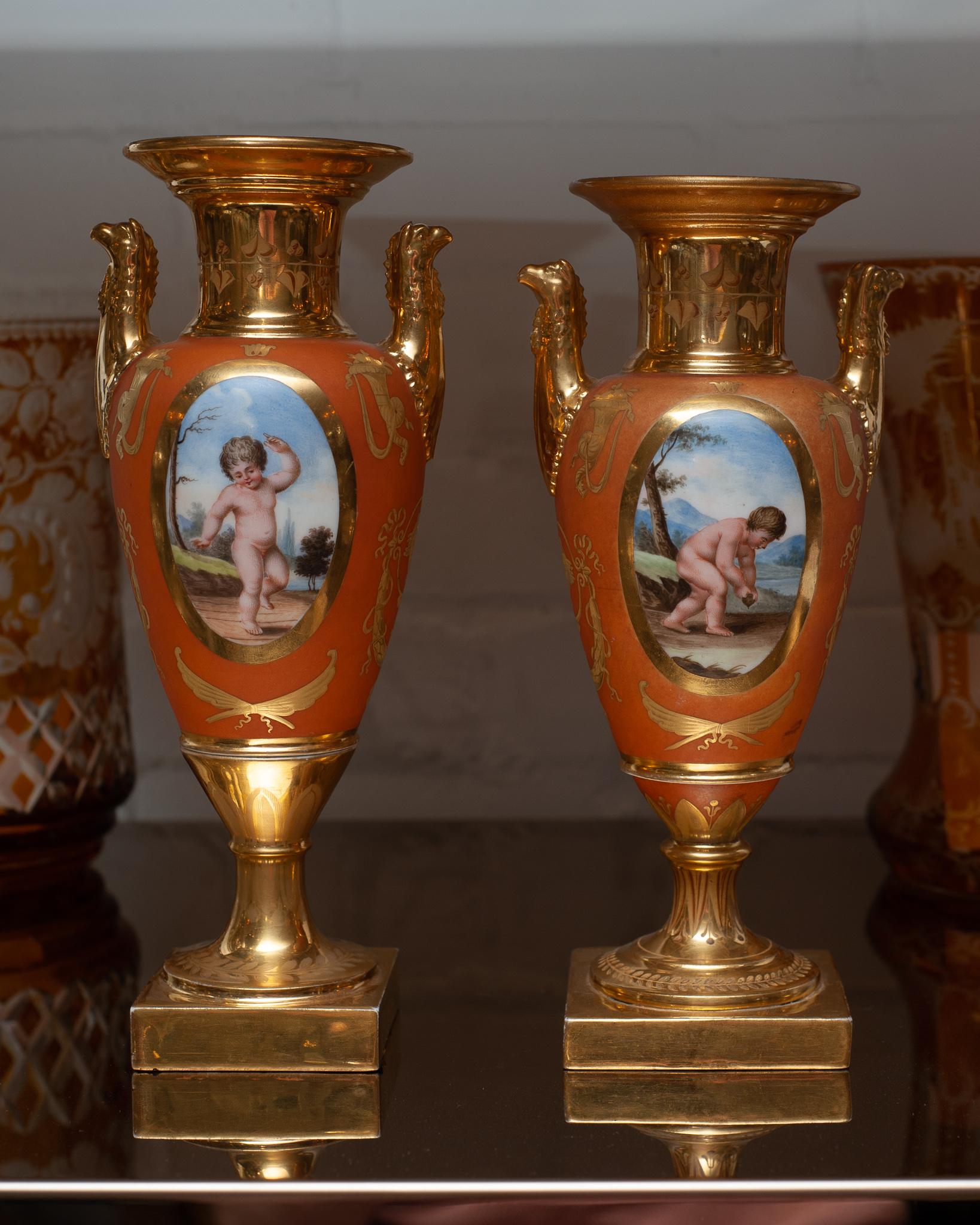 A beautiful pair of orange tone German hand painted urns, with gilded bases and detail. Each panel of either side of each urn depicts a unique painting. Gilded Griffin handles and pedestal bases.
