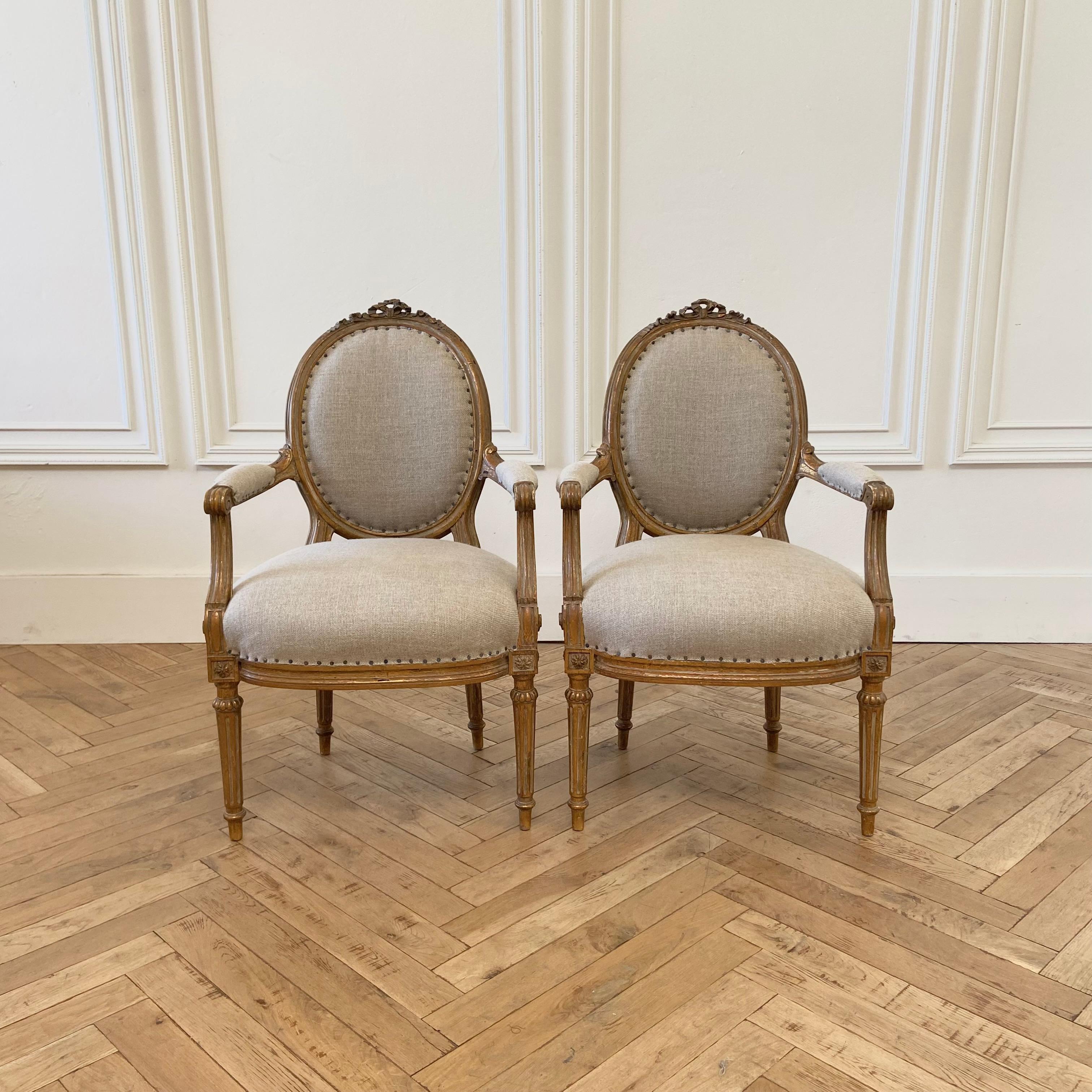 Antique pair of gilt wood open arm chairs upholstered in natural linen
Size:
23”W x 23”D x 36”H
Seat height: 19”
Seat depth: 19”
Arm height: 24”

These vintage chairs have the original finish, which is a weathered patina gilt wood. We have