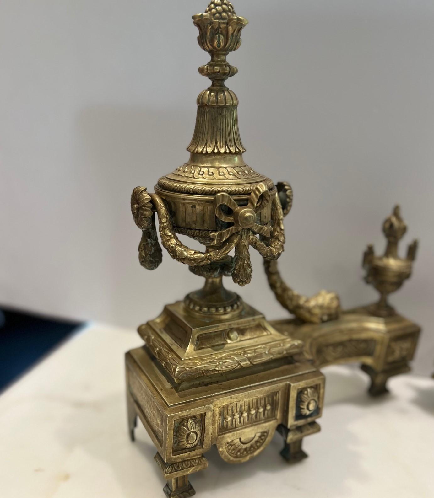Late 19th c. France, a pair of cast gilt bronze chenets/andirons modeled as a footed urn strung with a draping laurel and topped with a bowl of piled fruit. The larger urn is stationed beside a smaller flaming urn, both on a shaped plinth cast with