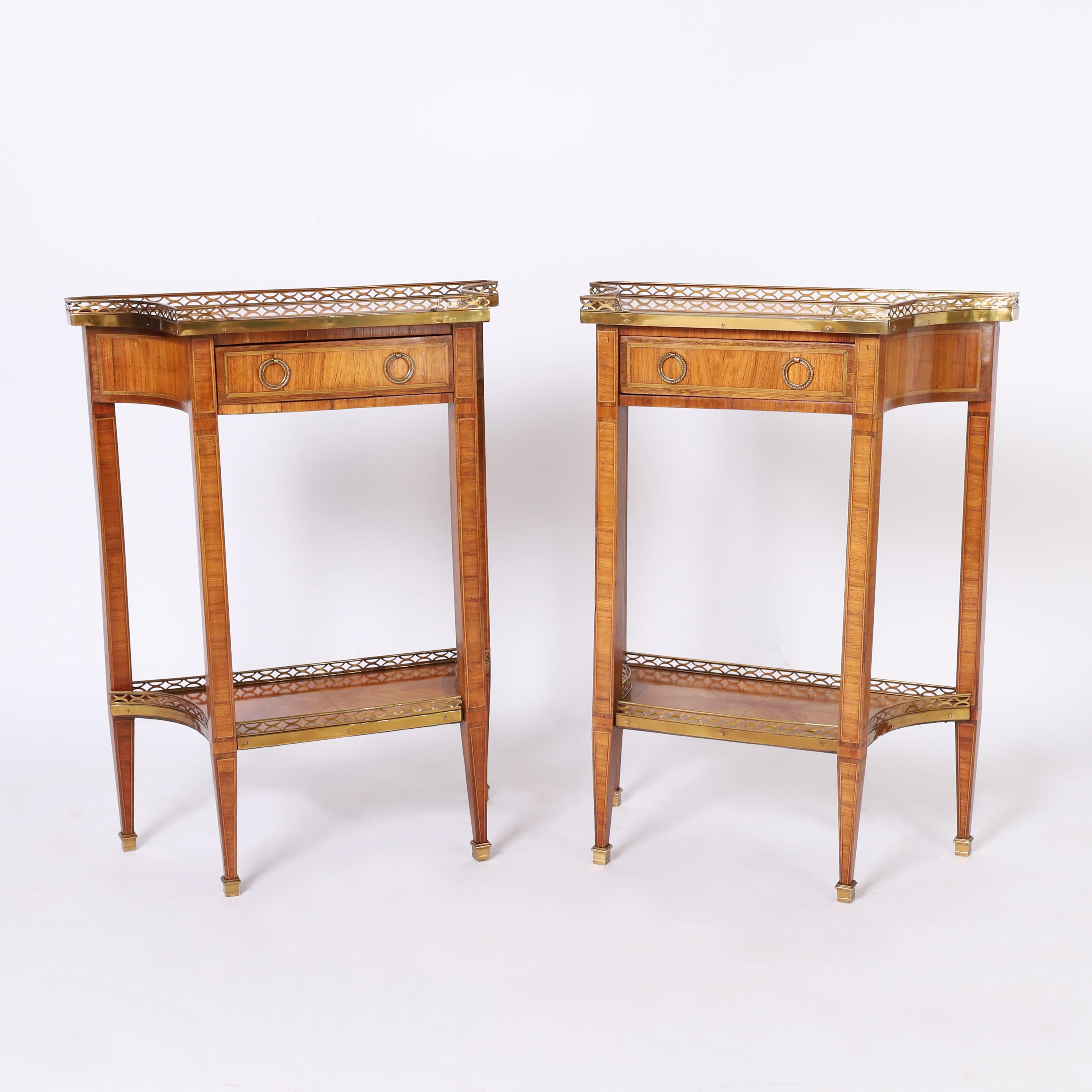 Standout pair of antique two tiered French stands crafted in satinwood in an unusual form with concave sides featuring marble tops, brass galleries, crossbanded inlays and tapered legs with brass spade feet.