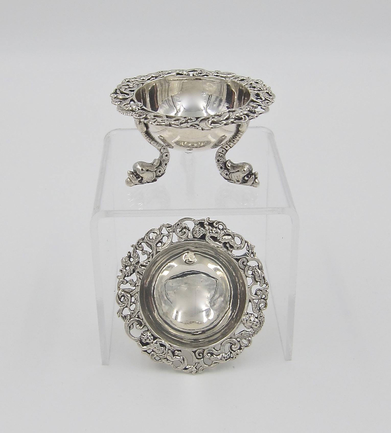 An antique pair of open salt cellars resting on three figural dolphin feet. Each salt is a simple bowl with a cast and pierced rim decorated with cherubs and scrolls. Each bowl is stamped on the underside with the letter 