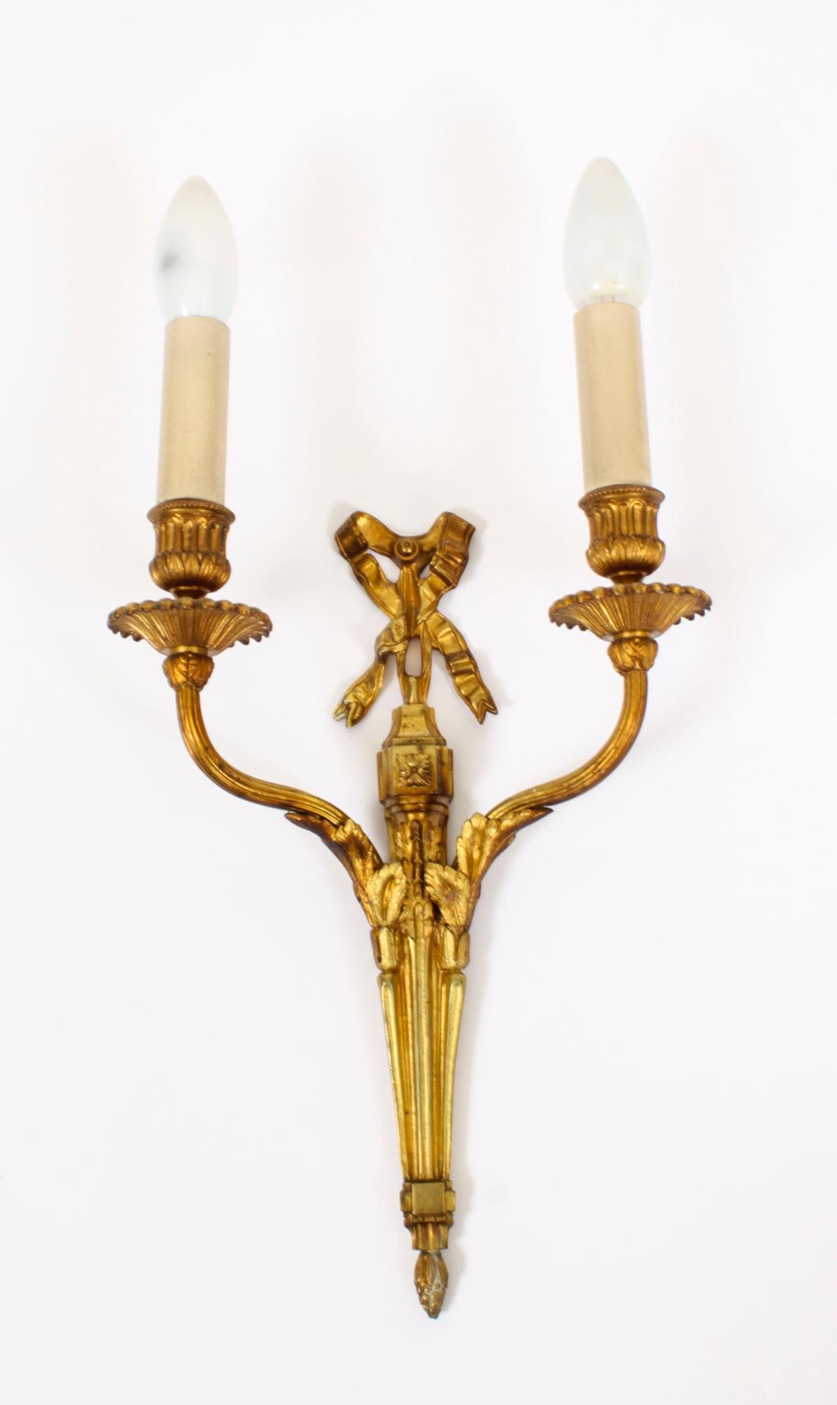 This is a fine pair of antique Regency Revival twin branch ormolu wall candle lights dating from Circa 1920.

The wall lights feature ribbon bow tops above tapering bodies with decorative mounts, each with acanthus leaf cast shaped branch arms with