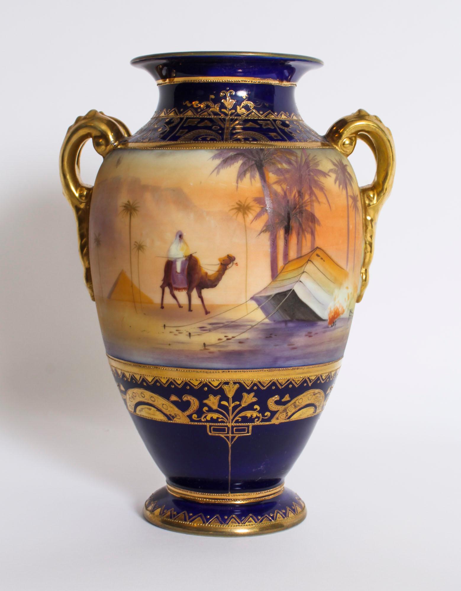 This is a beautiful antique pair of Taisho Period hand-painted twin handled Noritake porcelain vases, c.1920 in date.
 
They have an attractive and dominant navy-blue background with gilt high lights and feature painted scenes of figures and camels