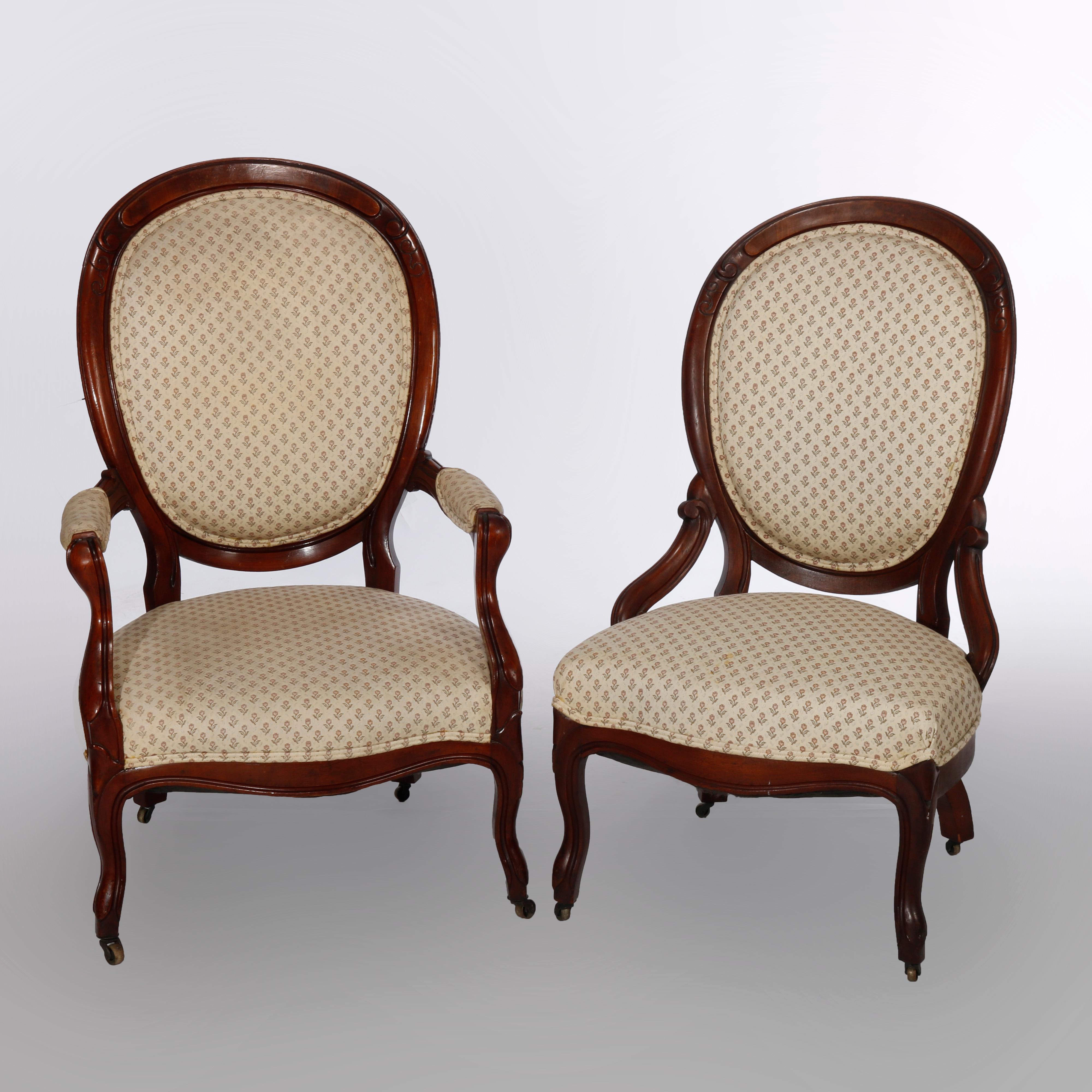 A pair of antique Victorian upholstered parlor chairs offer scroll form carved walnut frames with curved medallion form backs having burl insert rails, raised on cabriole legs and includes one gent's armchair and one lady's chair having skirt rails,