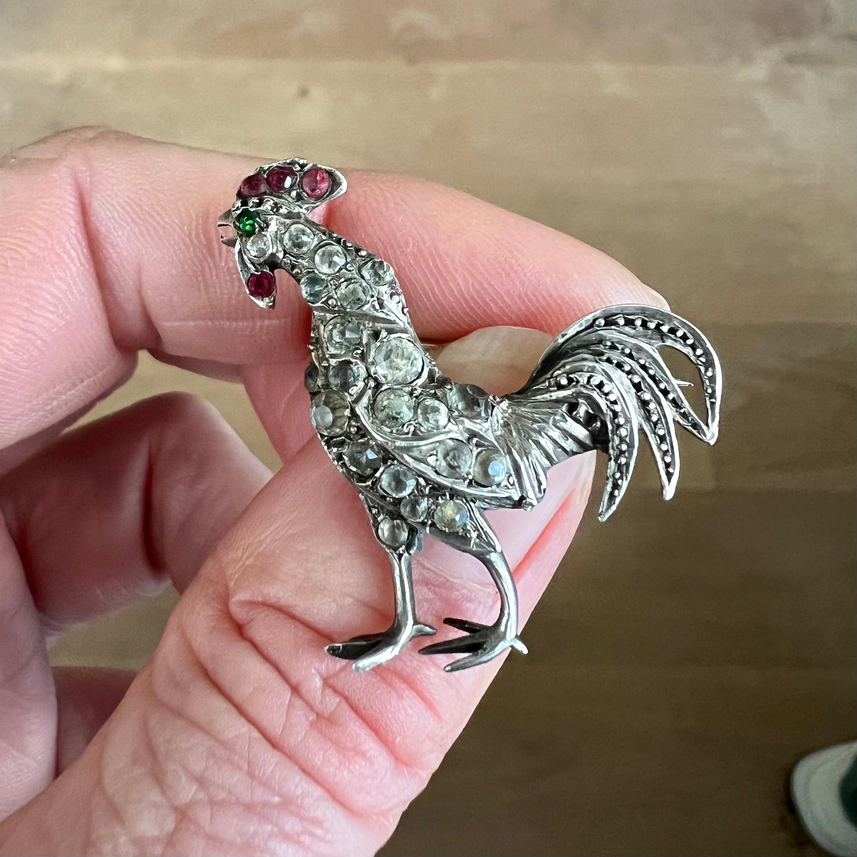 This is an antique silver rooster brooch set with translucent and colored paste glass stones. The rooster's body is set with diamond-like glass stones and his comb and wattle is set with red paste stones. The rooster's eye is made with a green paste