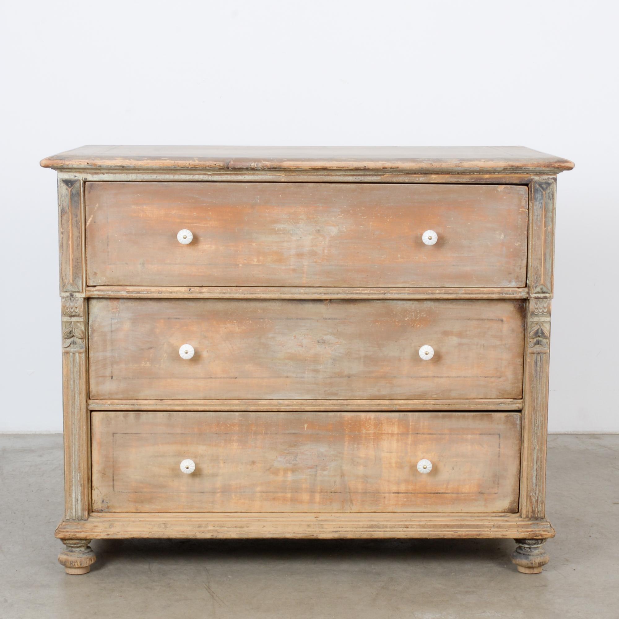 A wooden chest of drawers from Central Europe, circa 1900. The case is square; three ample drawers pull out with white ceramic knobs. The moldings are carved with distinctive designs, inspired by natural history — fluted columns evolve into