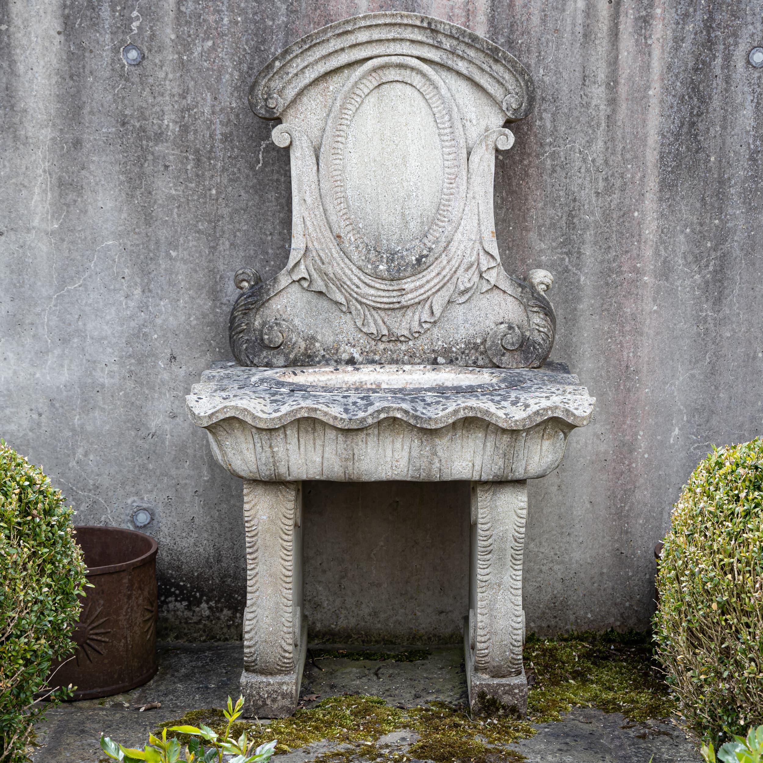 Fontaine murale en pierre naturelle patinée à l'ancienne avec panneau arrière profilé et bassin en forme de coquille. Des piliers de soutien solides et une construction robuste garantissent une stabilité durable.