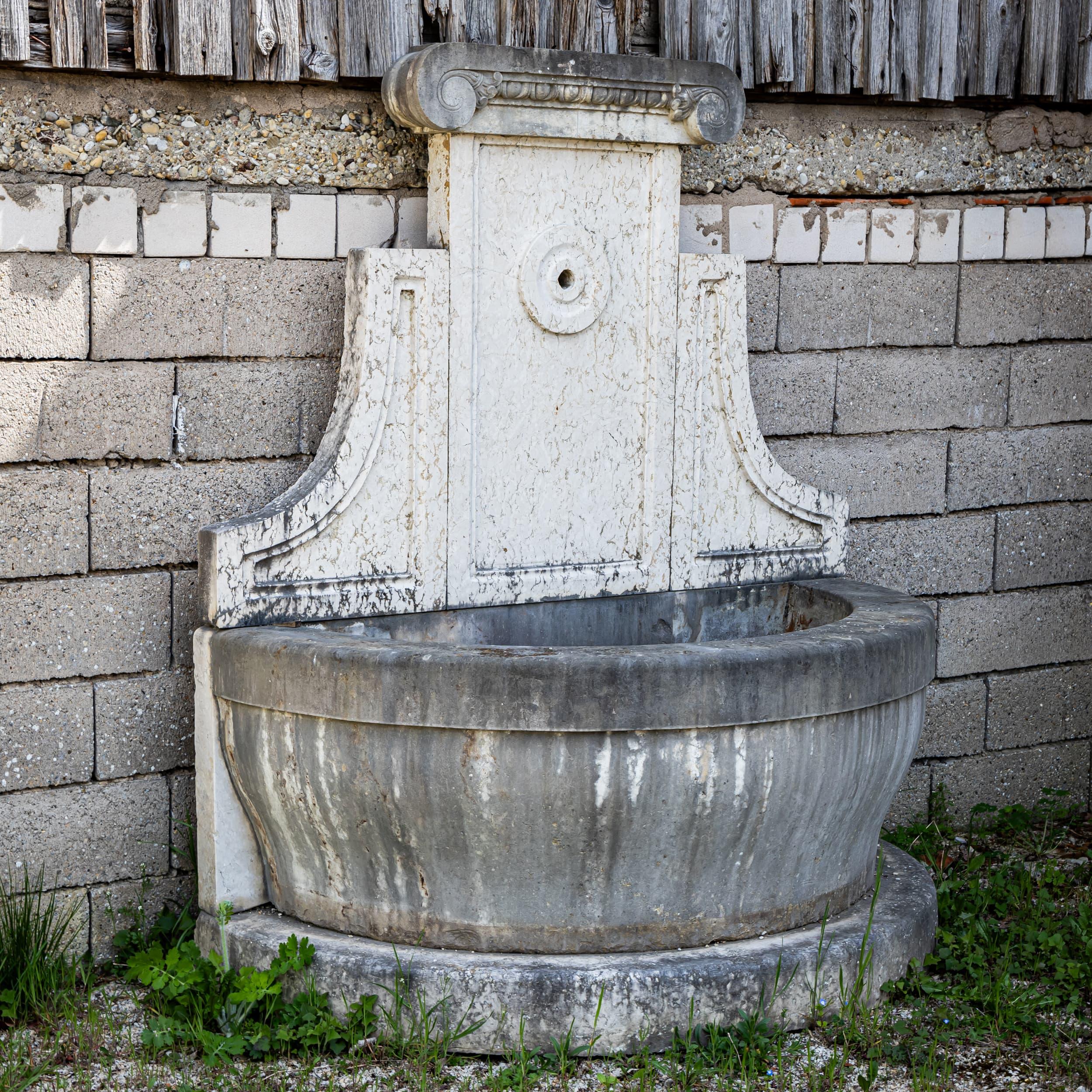 Wall fountain made of hand-chiseled, antique-patinated natural stone with a simple back panel and rolled top edge. The semi-circular, solid basin offers a harmonious shape and high stability.