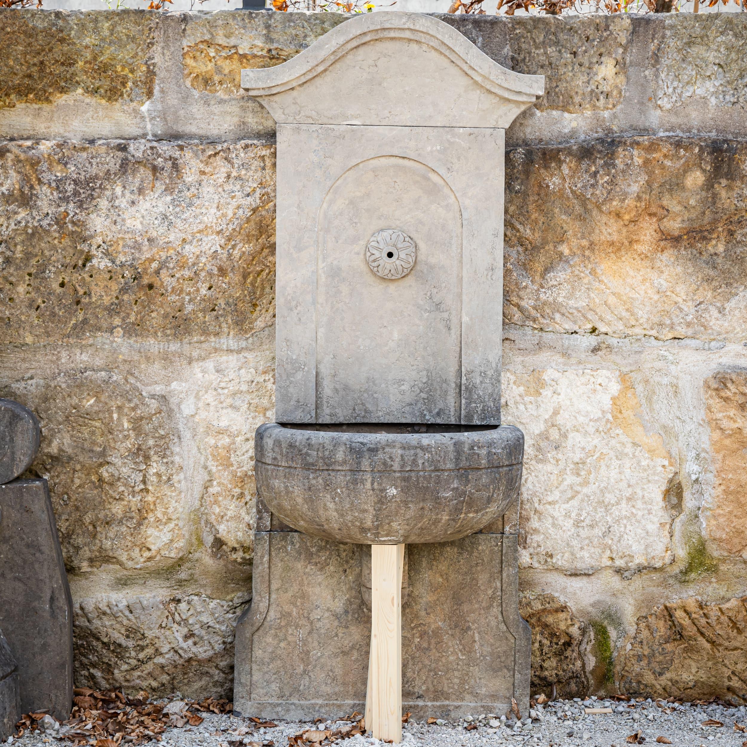 Fontana a muro in pietra naturale cesellata a mano e patinata all'antica con pannello posteriore curvo e beccuccio centrale a rosetta. Il bacino semicircolare e il design sottile offrono proporzioni eleganti e un'elevata stabilità.