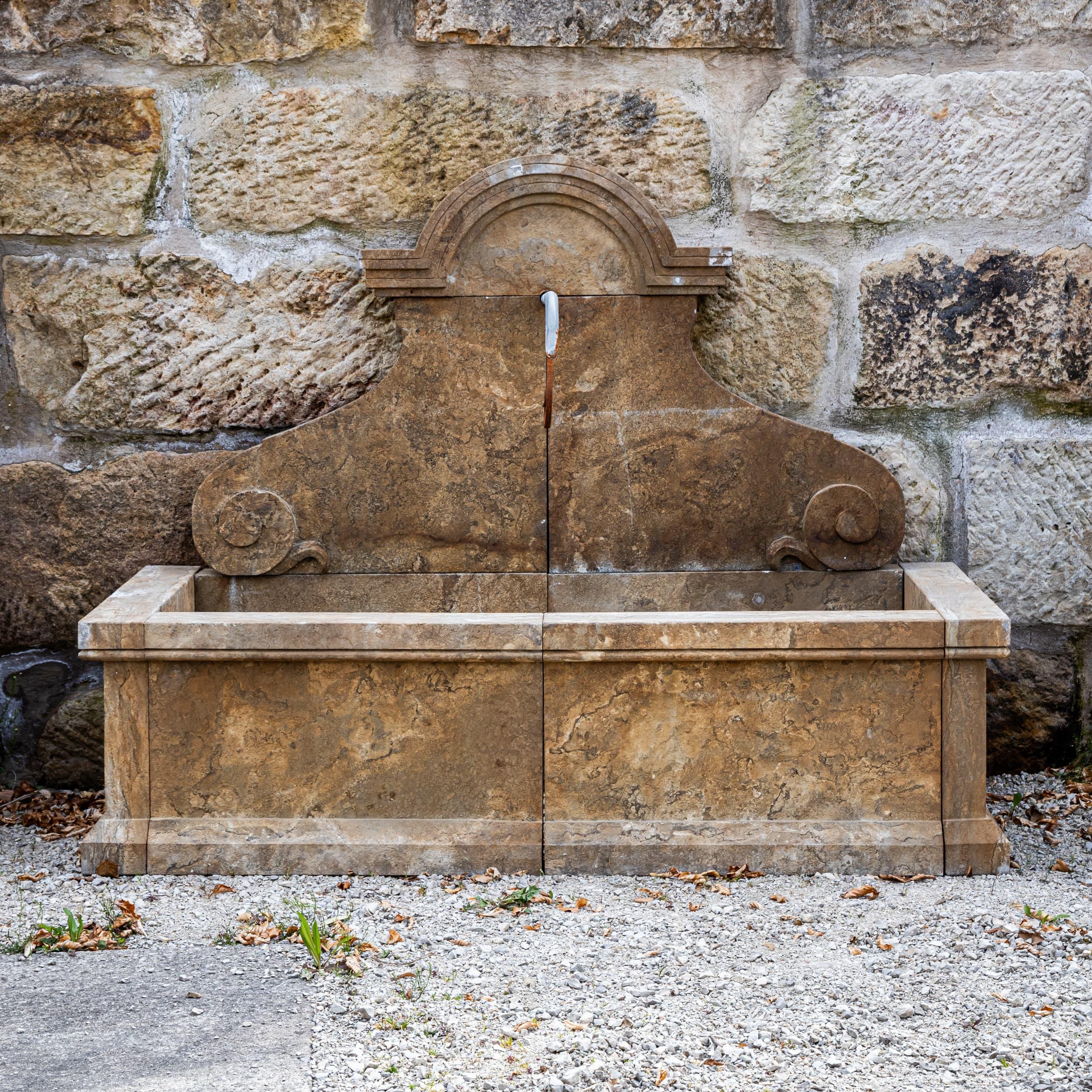 Wall fountain made of hand-chiseled, antique-patinated natural stone with a large rectangular basin. The curved back panel with subtle volutes and strong top molding gives the fountain classic elegance and high stability.