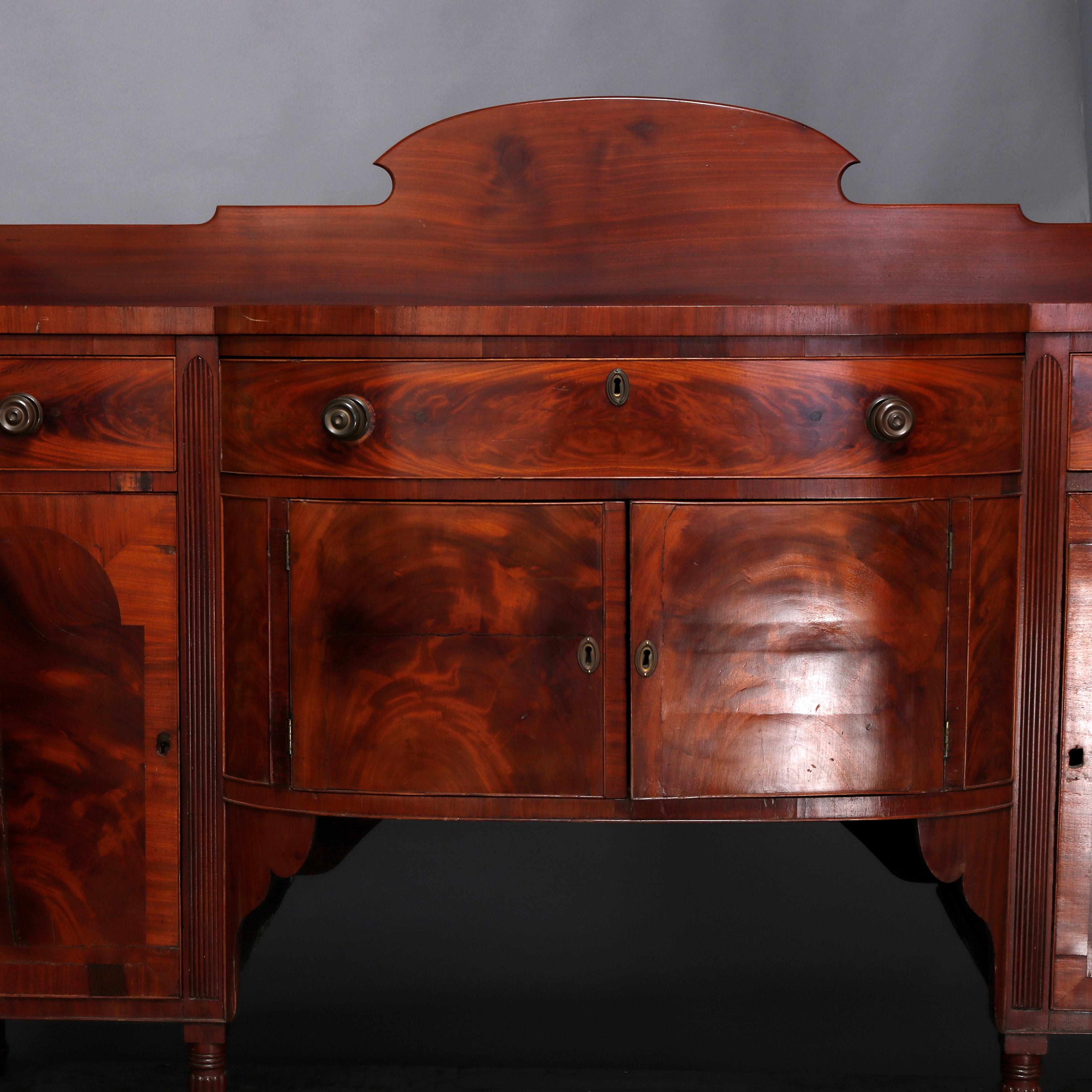 An antique period Sheraton sideboard offers flame mahogany construction with shaped backsplash surmounting case with bow front having frieze drawer flanked by two smaller and over double cross banded door cabinet flanked by two smaller, raised on