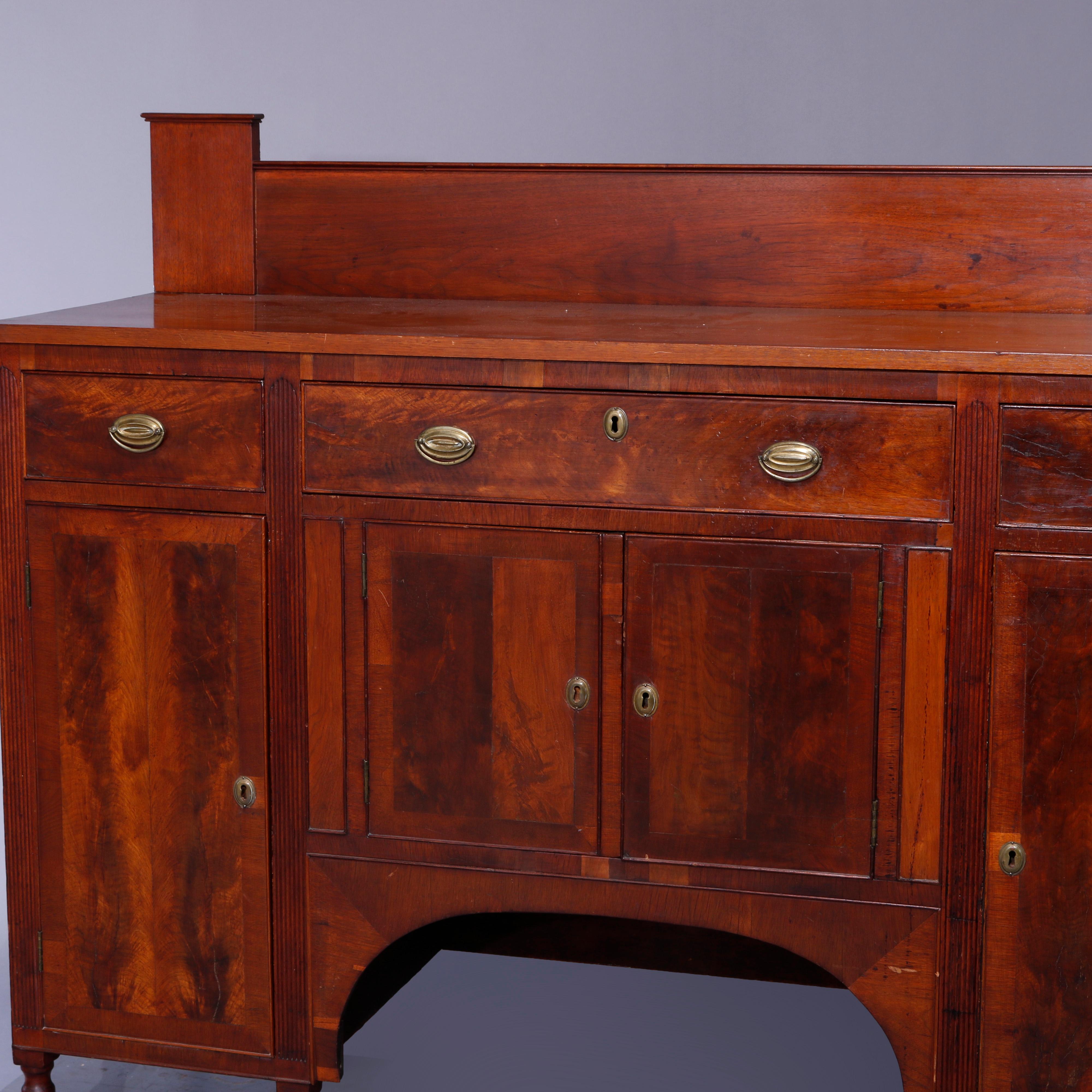 An antique period Sheraton sideboard offers flame mahogany construction with backsplash surmounting case with three upper drawers over lower cabinets having arched skirt, raised on turned legs, c1830

Measures: 52.5