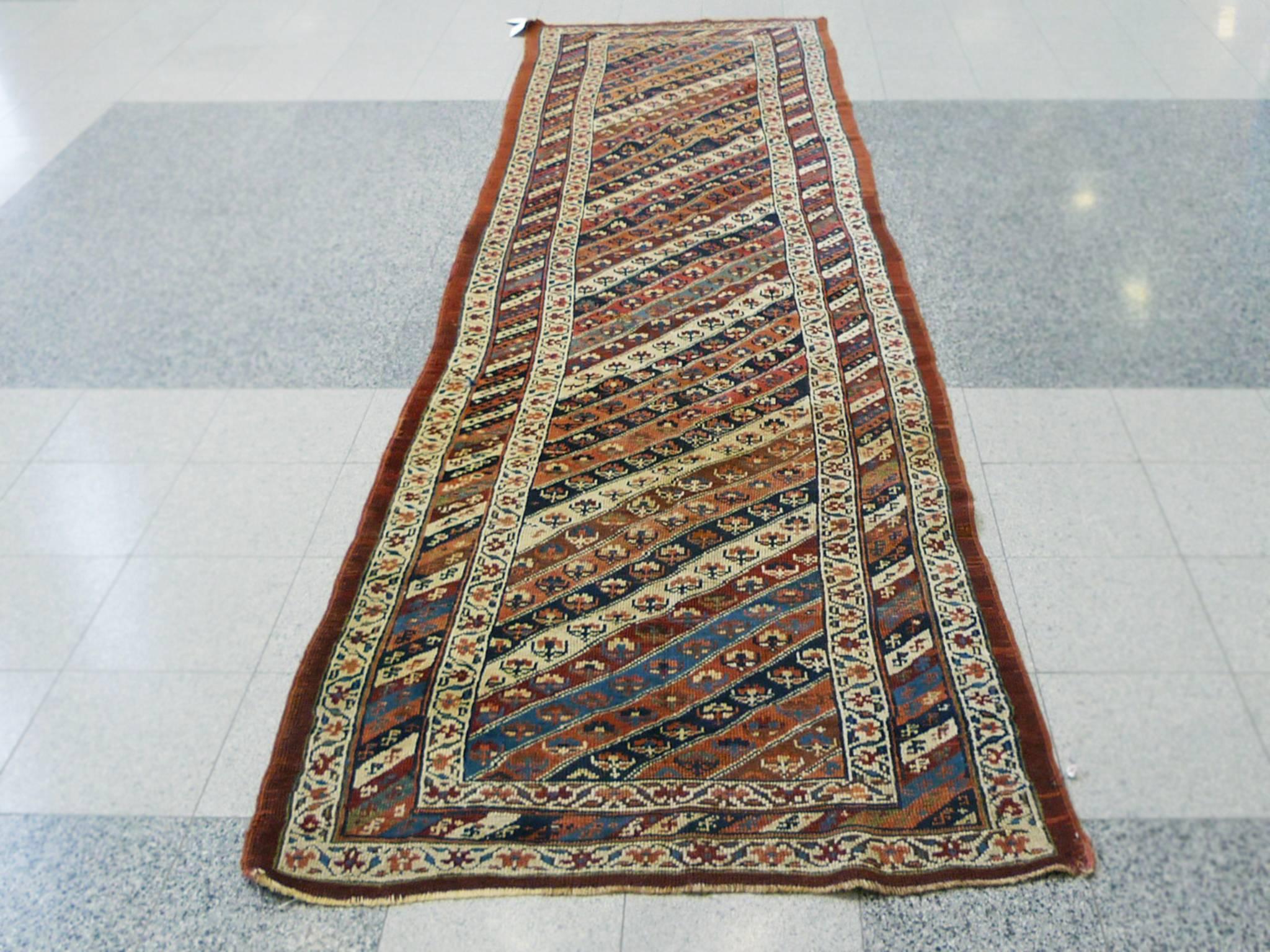 This richly patterned and textured runner rug is antique Persian Bidjar. It was handwoven in the Early 20th century. Its design consists of a central rectangle with diagonal stripes in yellow, pink, blue, and red hues. Surrounding this field is a