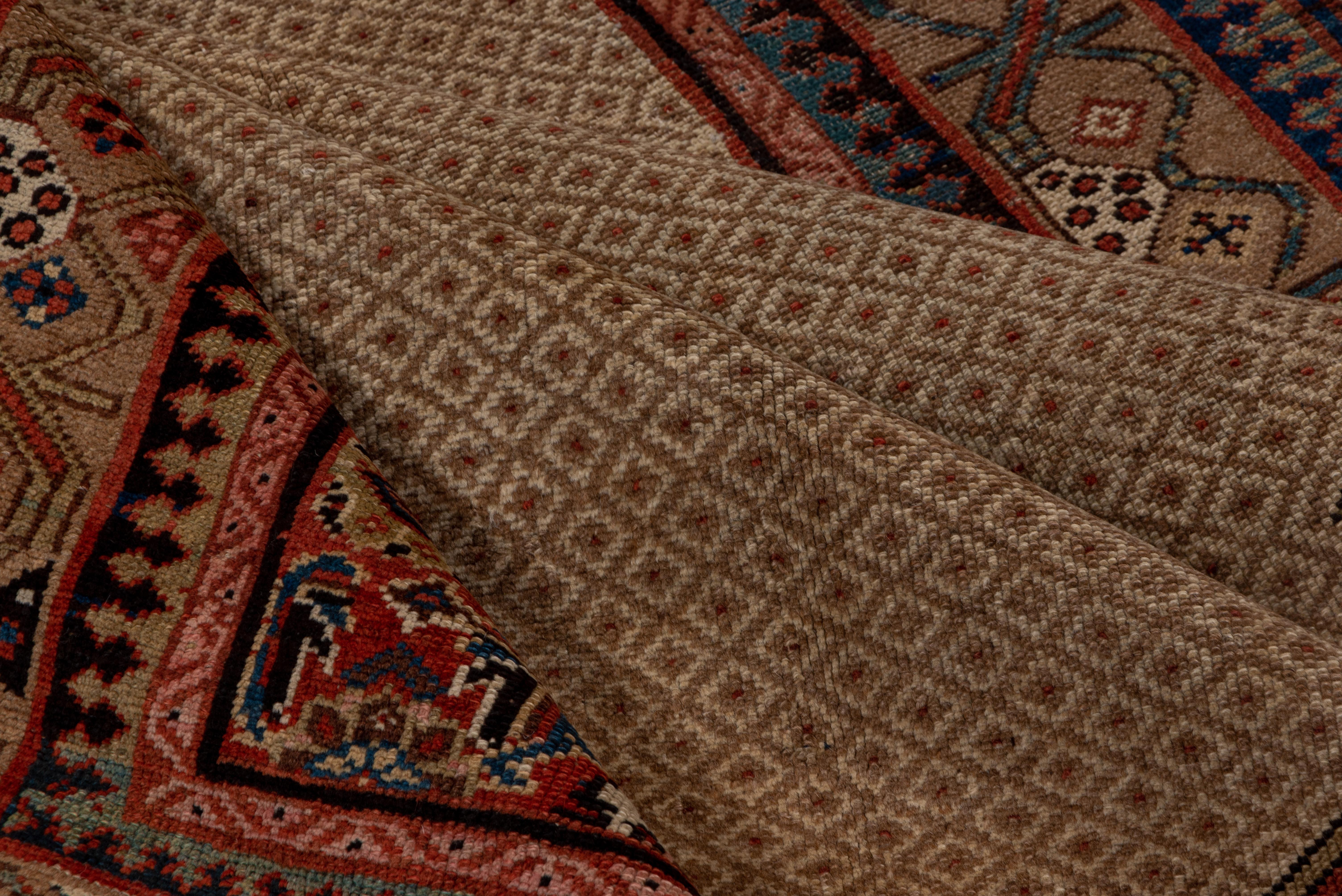 Le champ beige et camel à motifs minutieux accueille un médaillon Herati rouge central en forme de losange avec des coins en suite, avec une bordure principale de couleur camel composée de feuilles obliques en barre et de têtes de fleurs