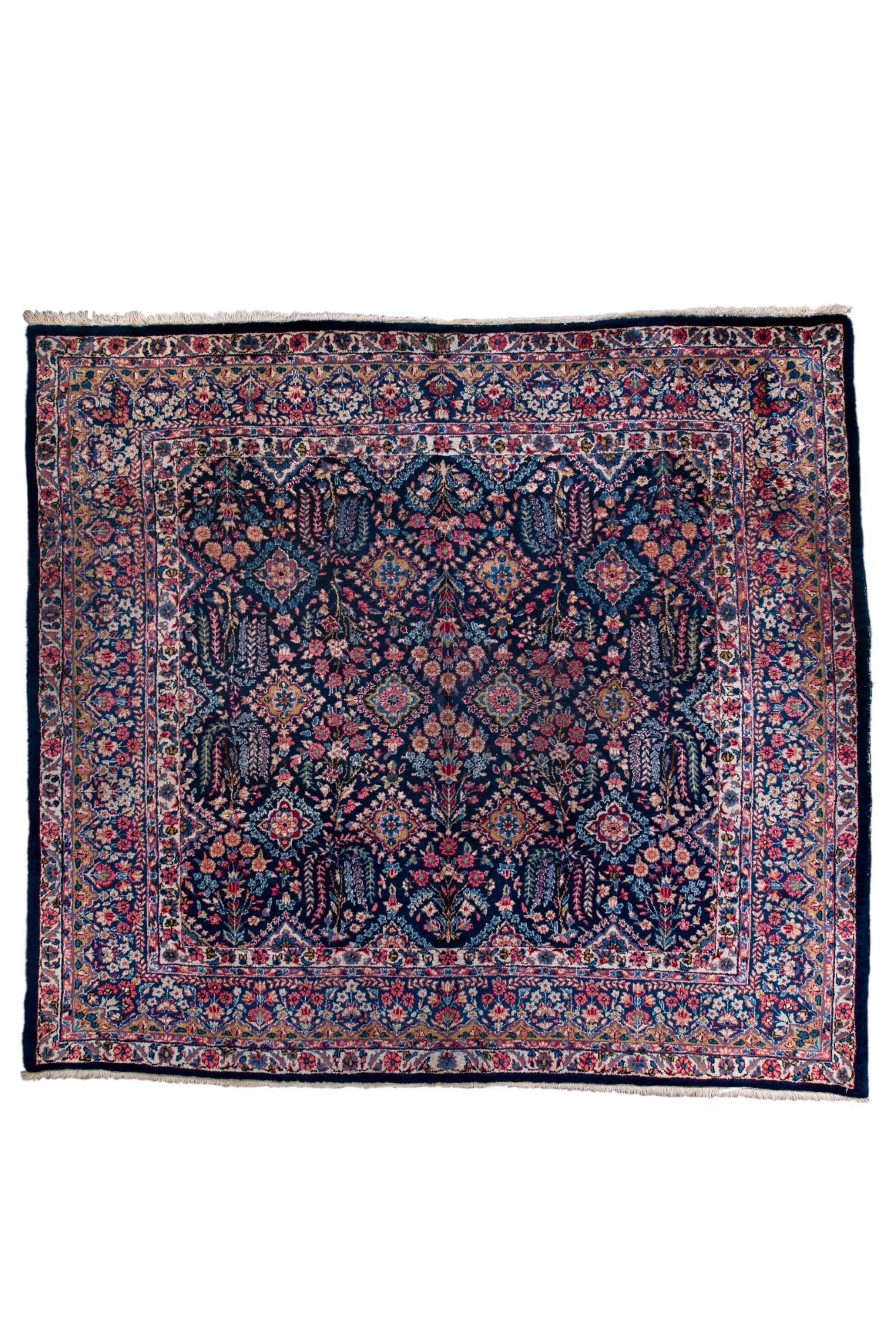 This antique Kerman rug features a dense allover floral medallion pattern set on a deep navy field, framed by classic Kerman borders filled with scrolling blossoms. The refined palette—navy, rose, ivory, and soft greens—highlights the region’s