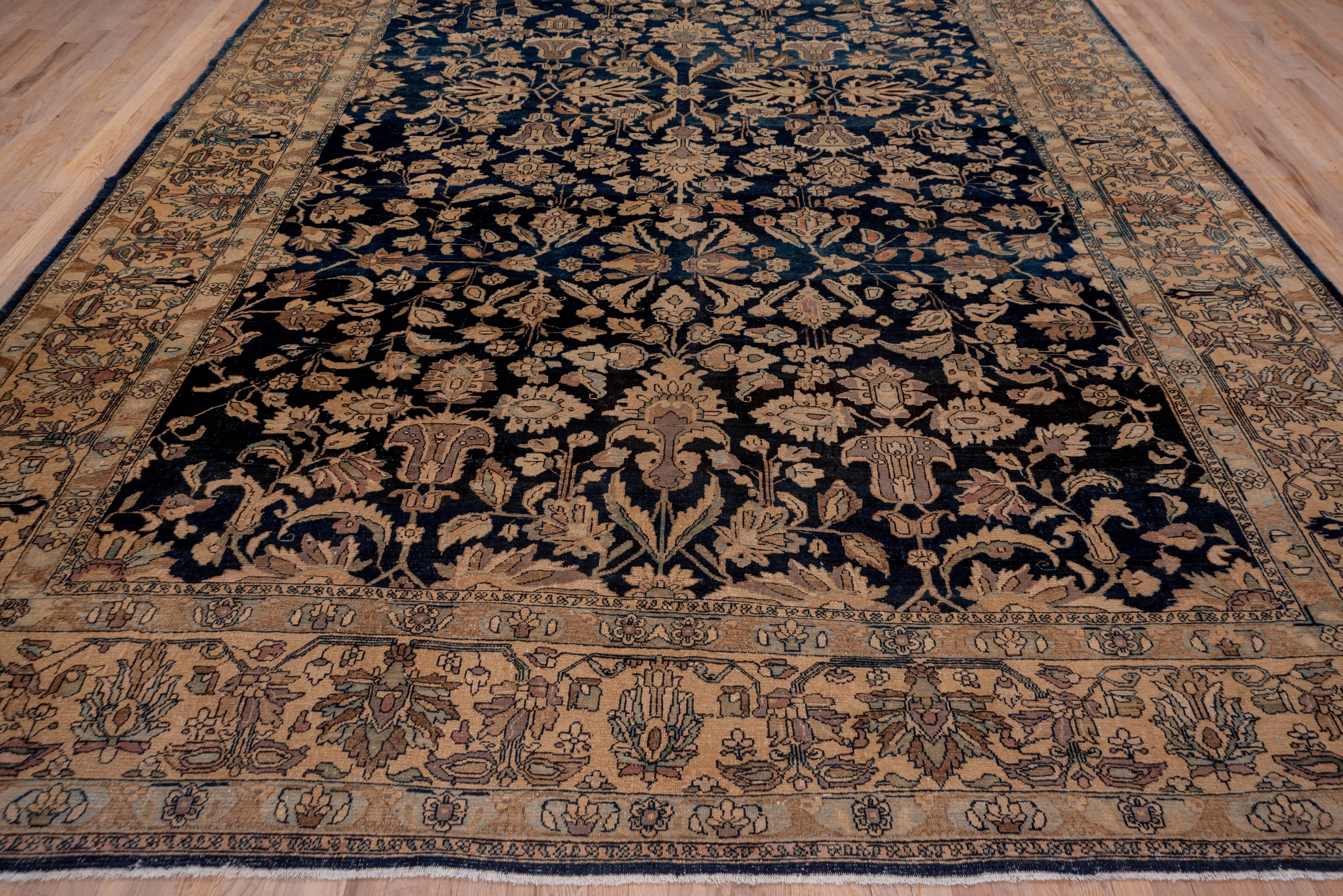 This west Persian town carpet shows a navy blue ground with an all-over escutcheon, palmette, blossom and leaf pattern. The beige border displays two varieties of in and out palmettes sprouting small flowers. The light toned border system is a nice