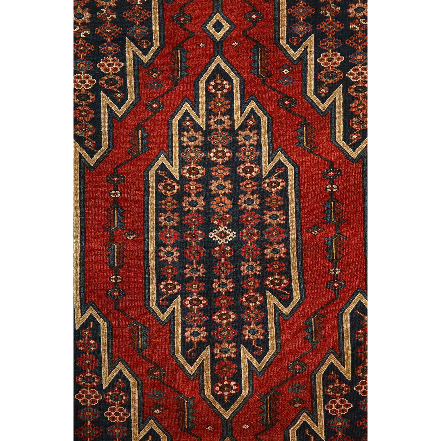 This antique Persian Mazlaghan carpet in pure wool and vegetable dyes circa 1920 features a geometrically striking central medallion with layered borders and a patterned field of stylized floral motifs. Its bold patterning is balanced by its organic