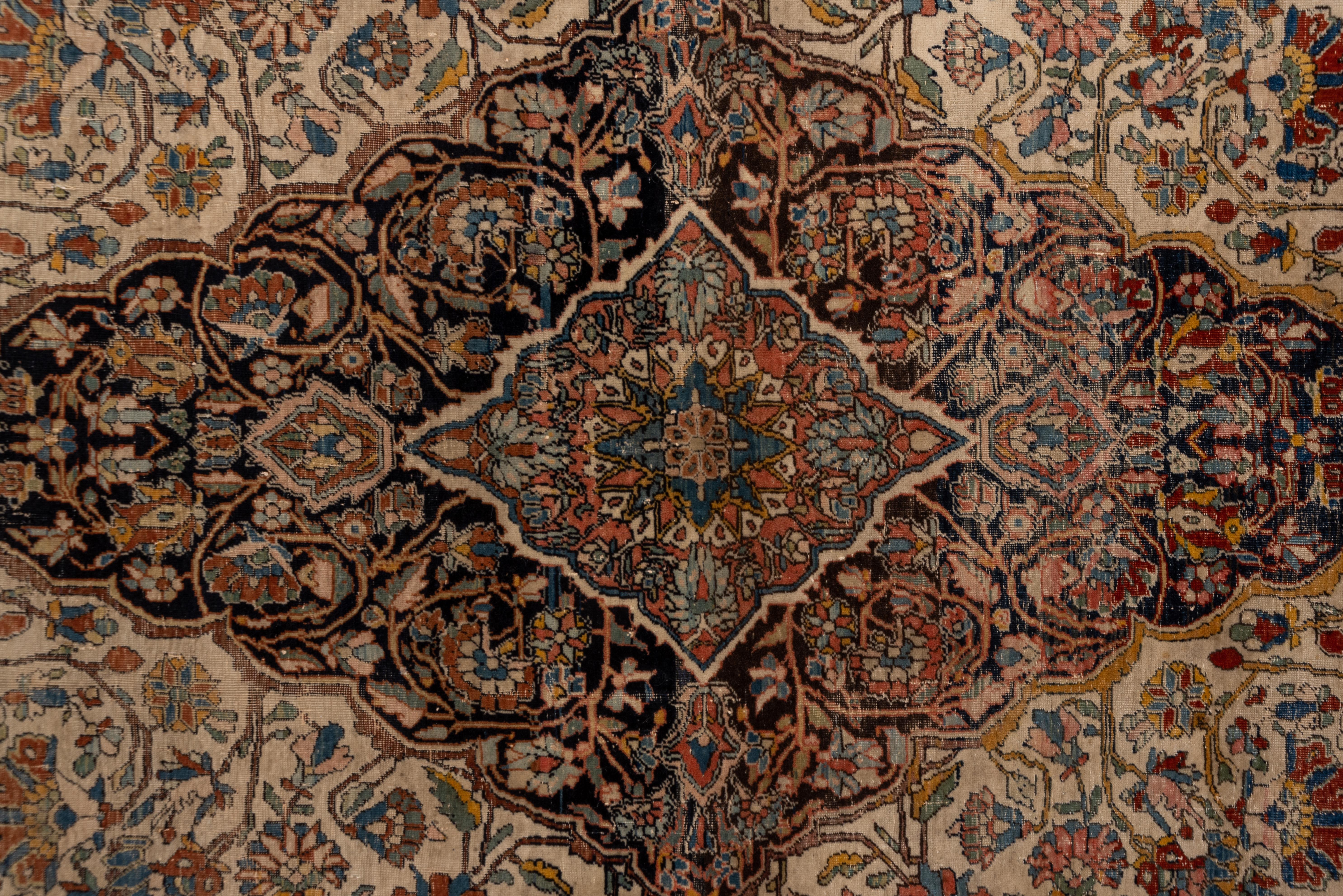 This antique, finely woven, central Persian city scatter rug shows a scalloped, lightly abrashed navy palmette medallion centering a rose Sub-medallion, all on a cream field with en suite decor. The cream border shows large, petal rosettes and