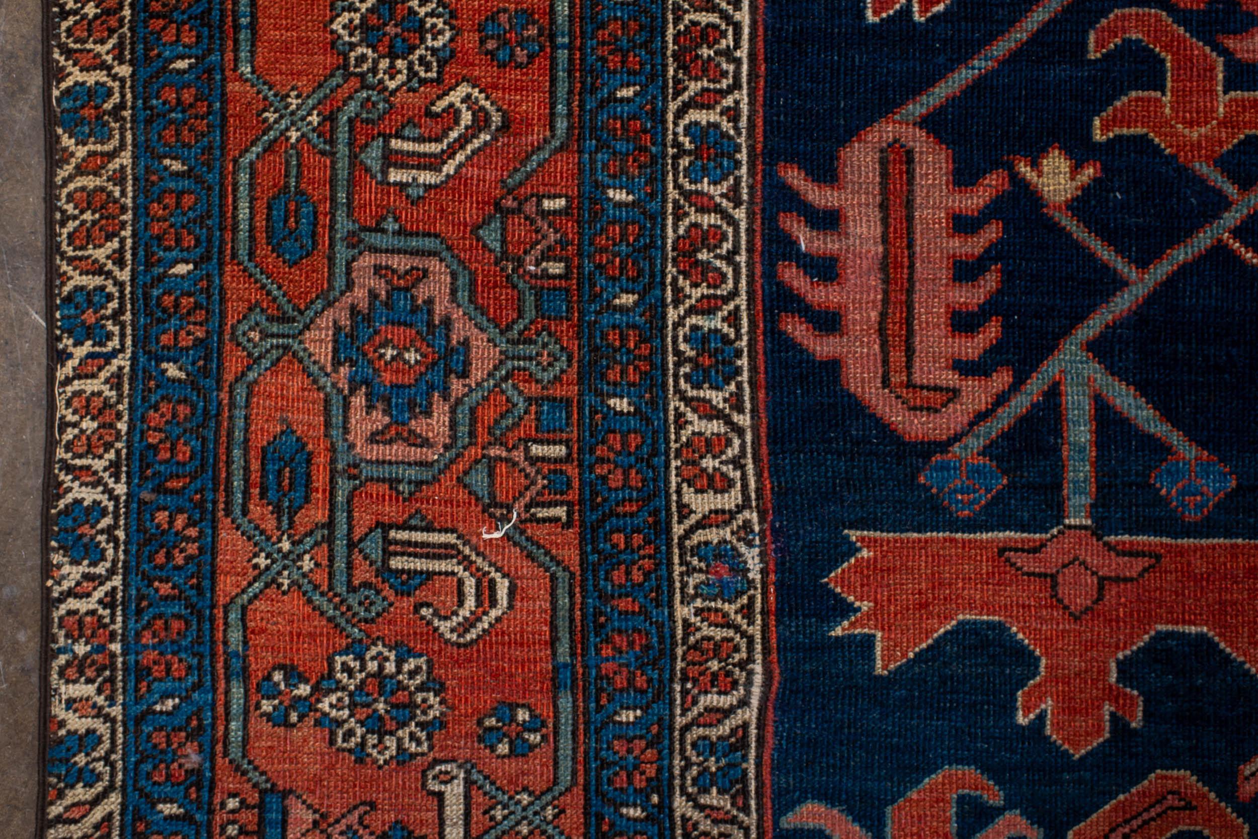 Un tapis traditionnel Heriz de conception géométrique avec un fond bleu marine profond entourant un motif central ivoire et rouille, les écoinçons soulignant le motif général. Les bordures principales et secondaires qui l'entourent contribuent à