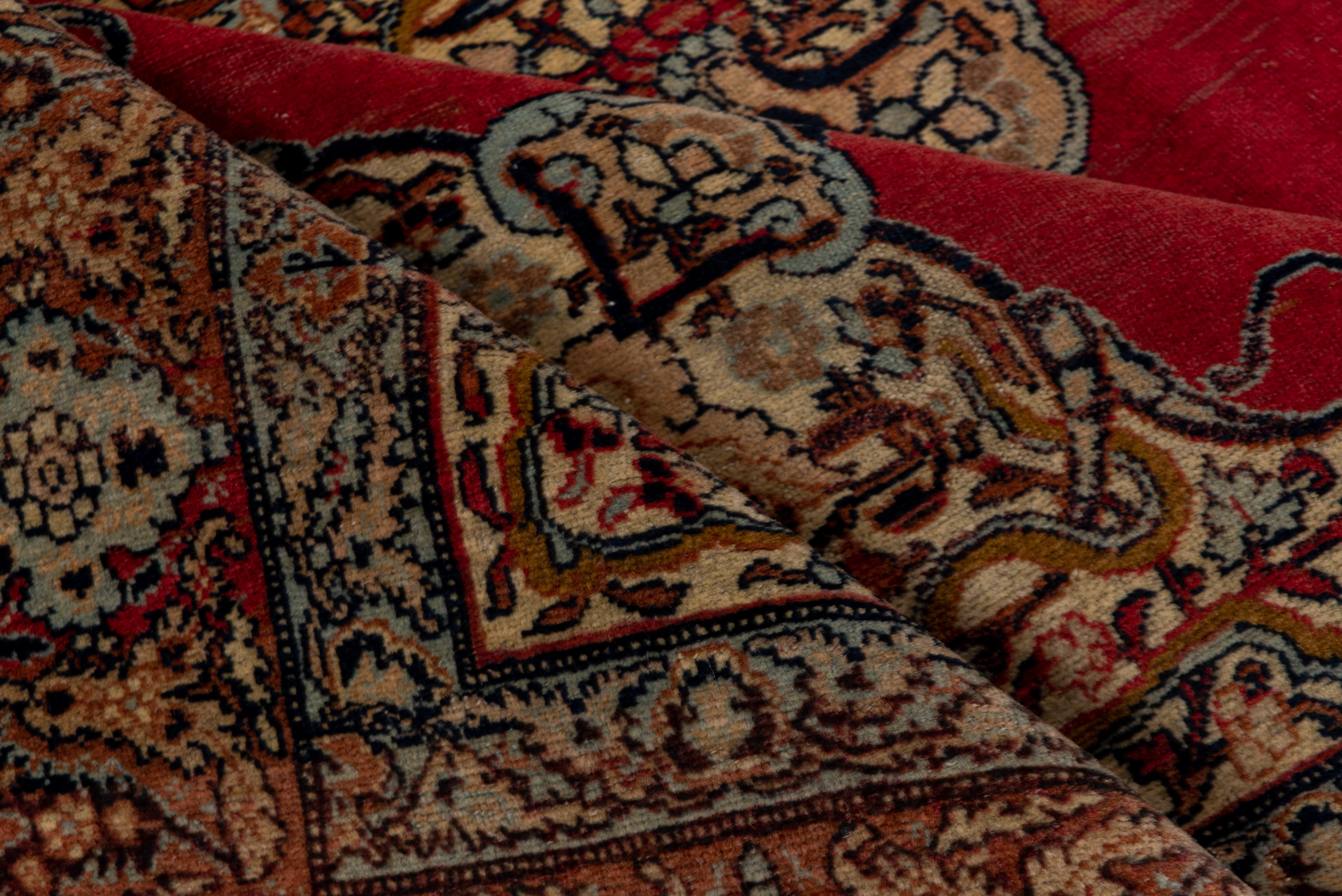 This antique Tabriz rug has a sand-straw four pendant medallion with an inner decor of tightly layered arabesques and cloud bands is set on the open ruby red field within en suite corners with more snaky cloud bands. Salmon-pink border of softly
