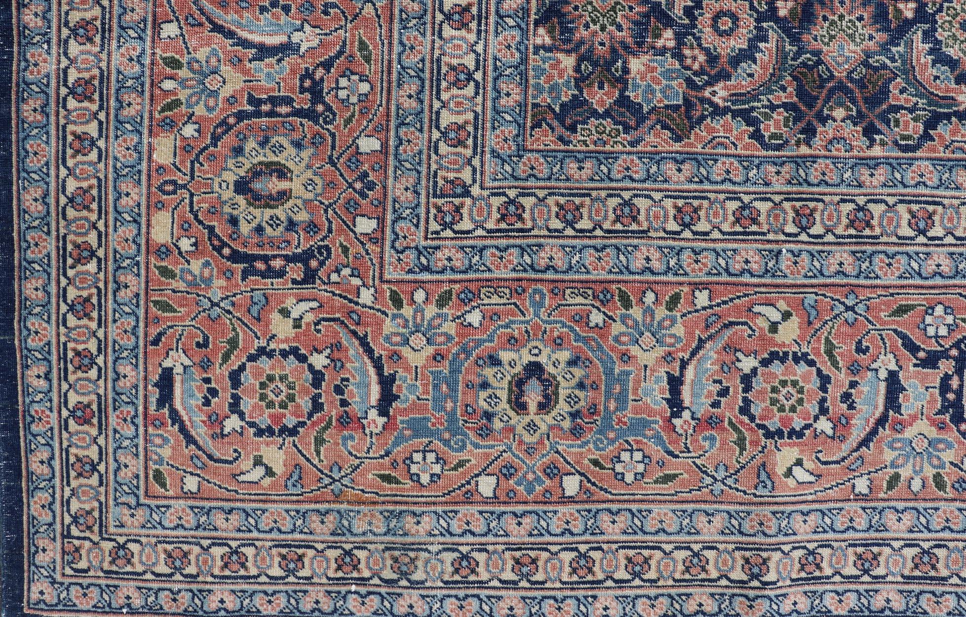 This hand knotted antique Persian Tabriz rug features an all-over, sub-geometric floral design rendered in blue, coral, orange, ivory and blue shades, set upon a navy blue background. A complementary, multi-tiered border encompasses the entirety of